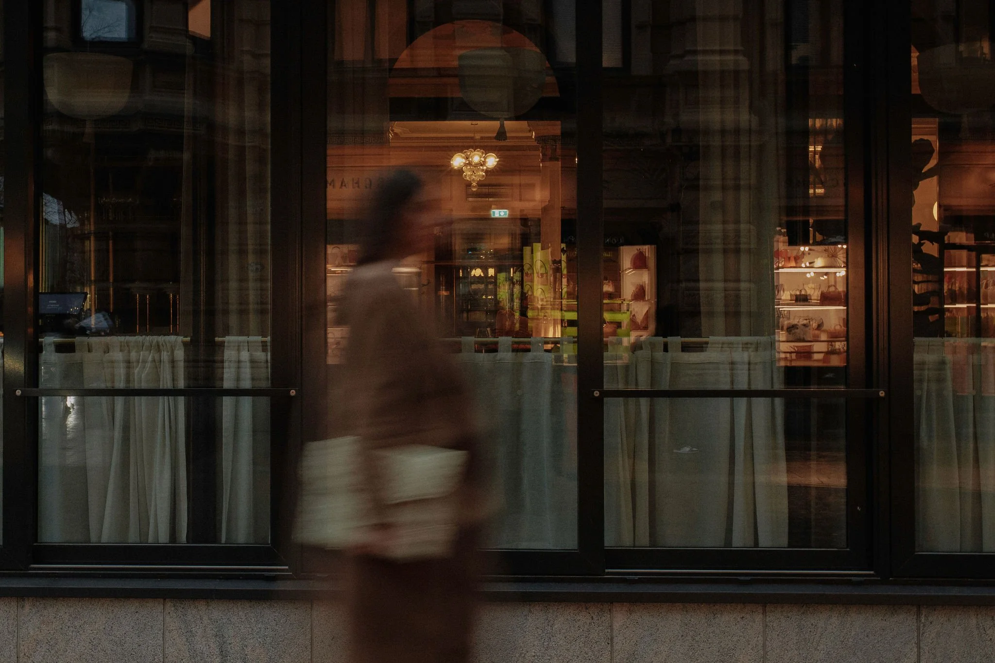 A blurry woman walking past a glass window of a restaurant or cafe, showing interior lighting, decor, and shelves inside.