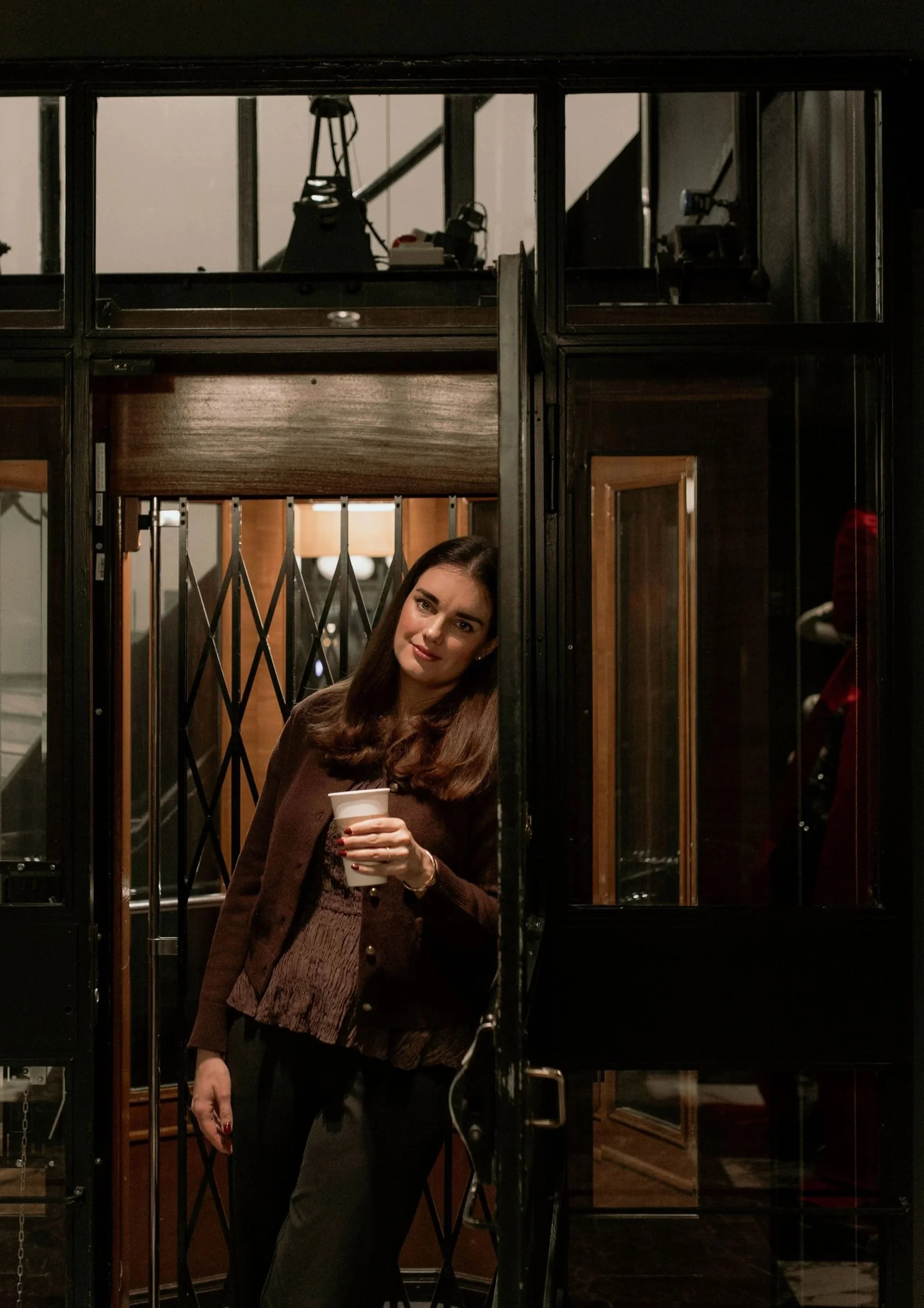 A woman with dark hair, wearing a brown jacket, holding a white cup, stands inside a guarded elevator in a dimly lit indoor setting.