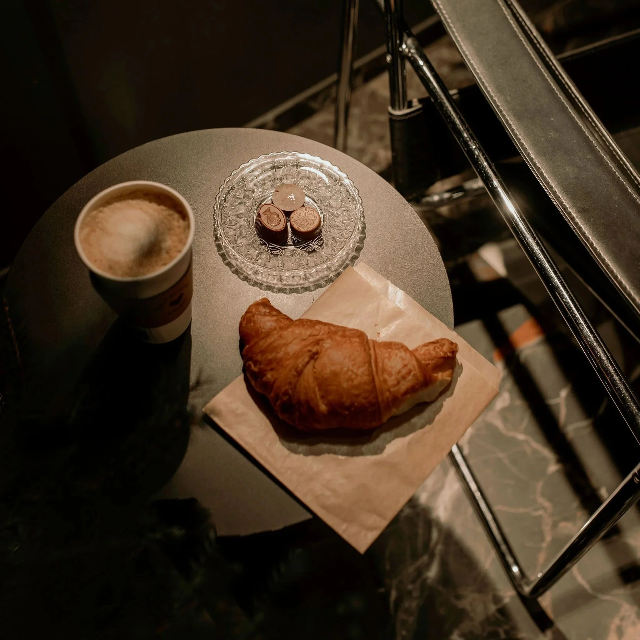 A table with a cup of coffee, a croissant on parchment paper, a glass plate with chocolates, and a stack of metal chairs in a dimly lit setting.
