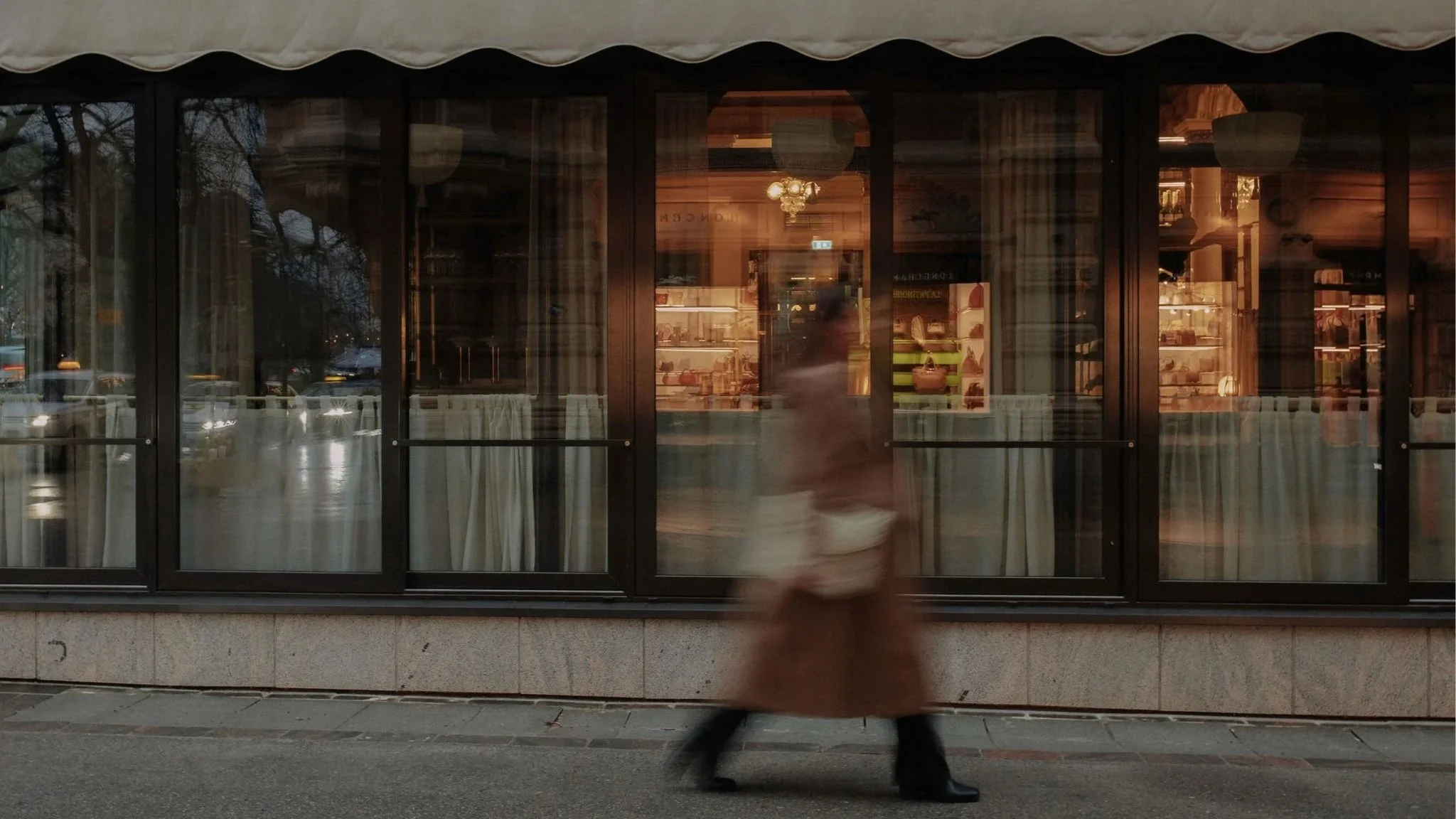 Blurry person walking past a storefront window with curtains and illuminated interior display, trees and cars visible in the background.