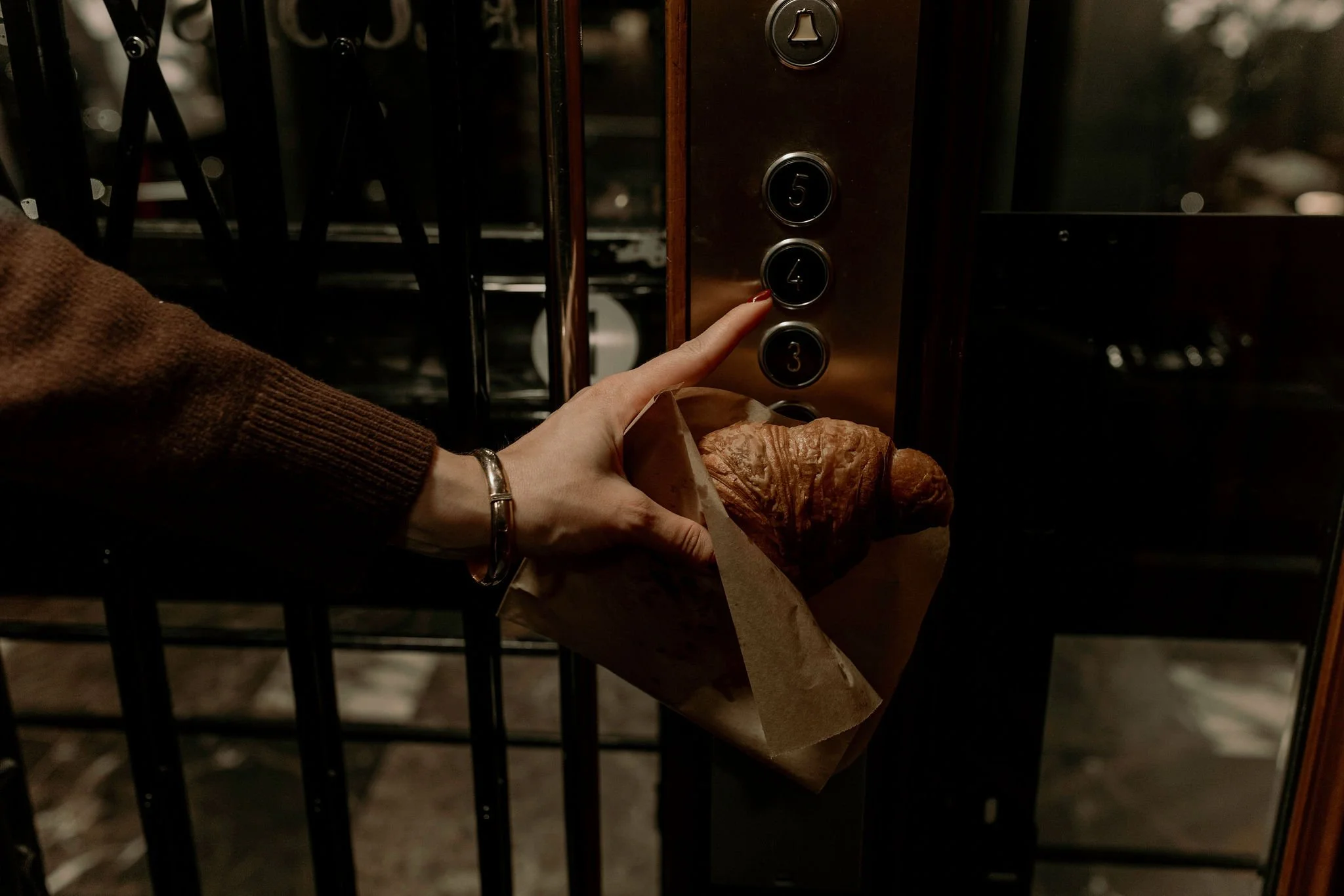Person pressing the elevator button labeled '4' while holding a croissant in a brown paper bag.