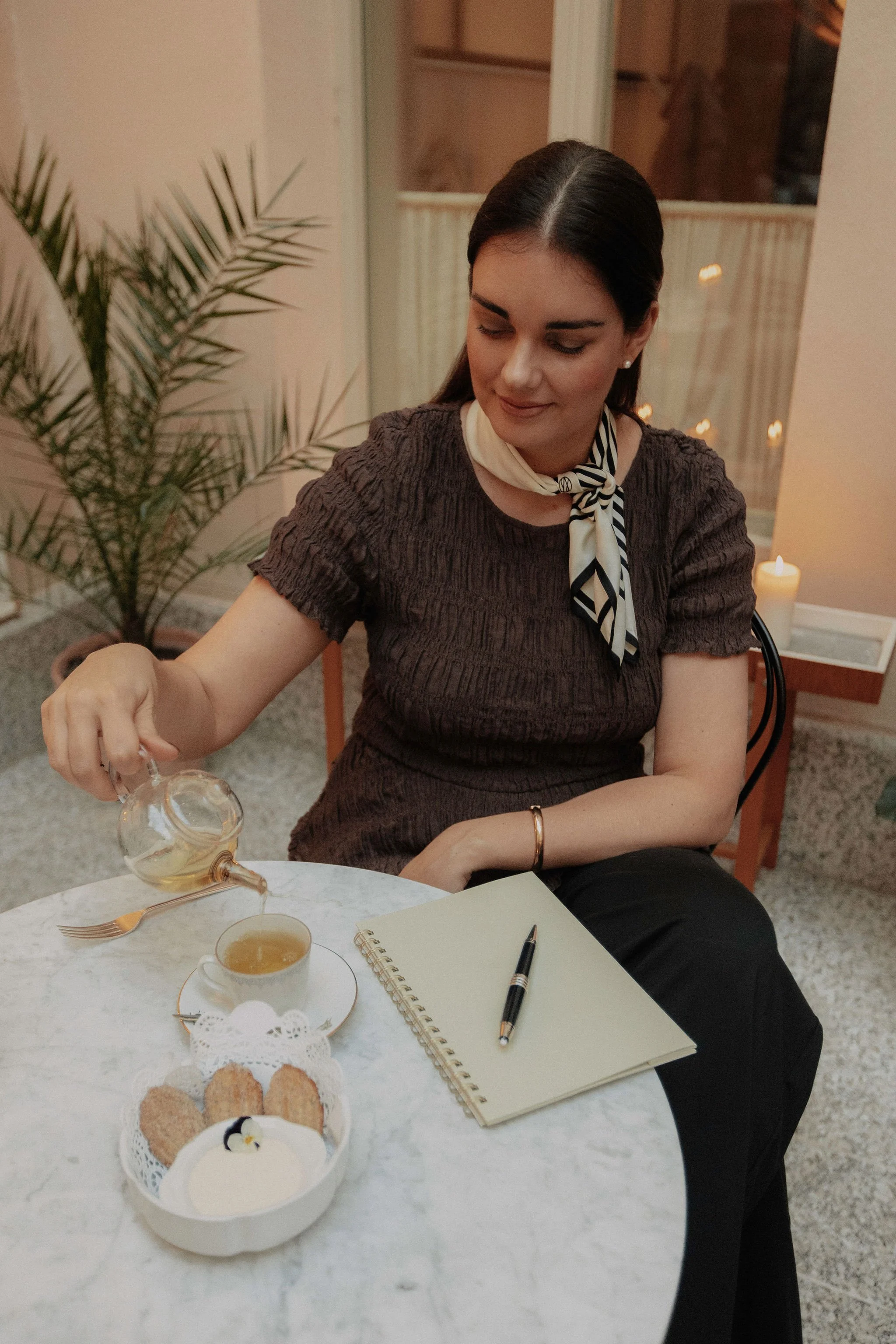 A woman with dark hair tied back, wearing a brown top and a black and white striped scarf, is sitting at a marble table pouring tea into a cup.