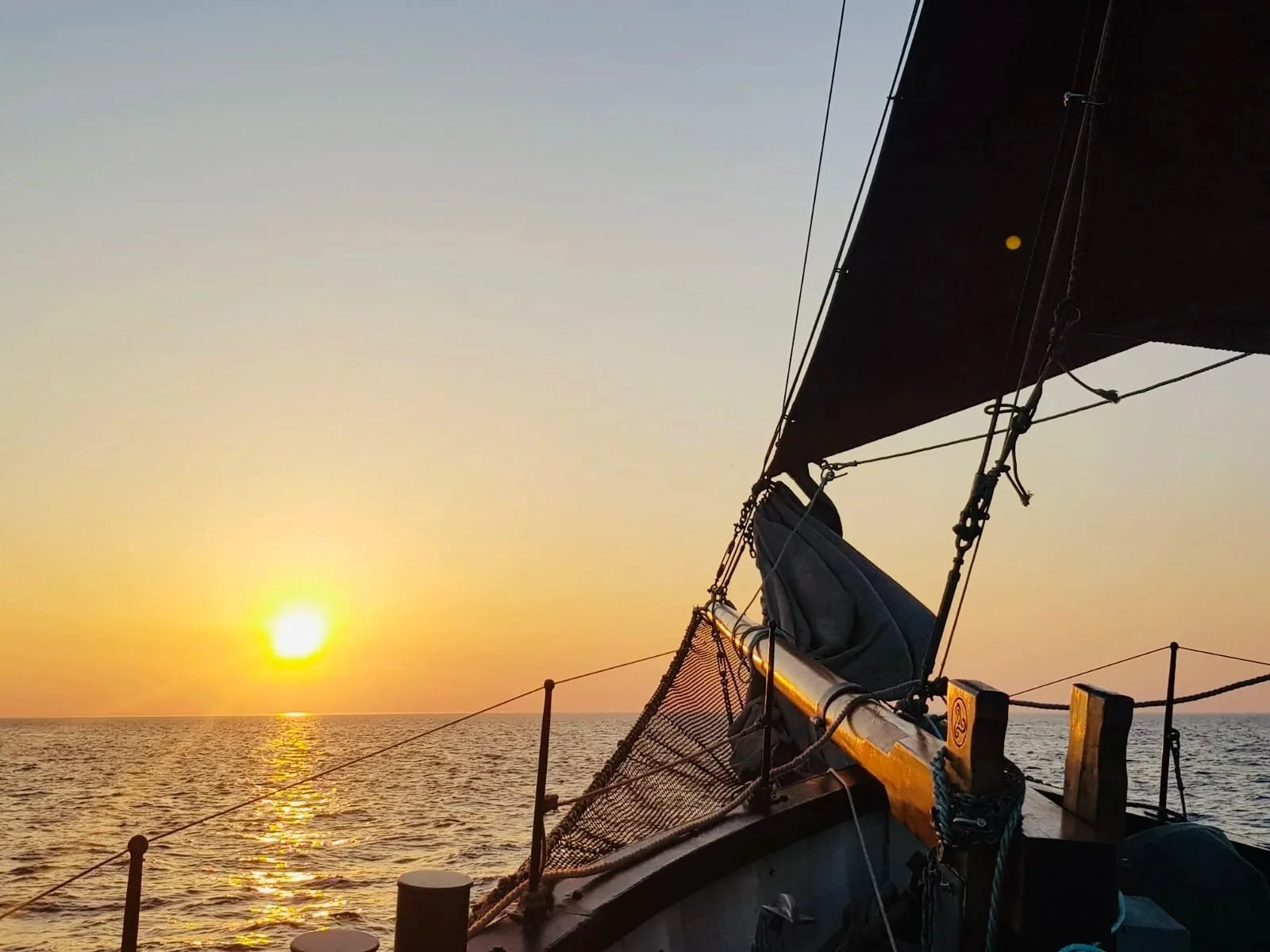 View of a sailboat deck at sunset, with the sun near the horizon over calm water, and the boat's mast and rigging visible.