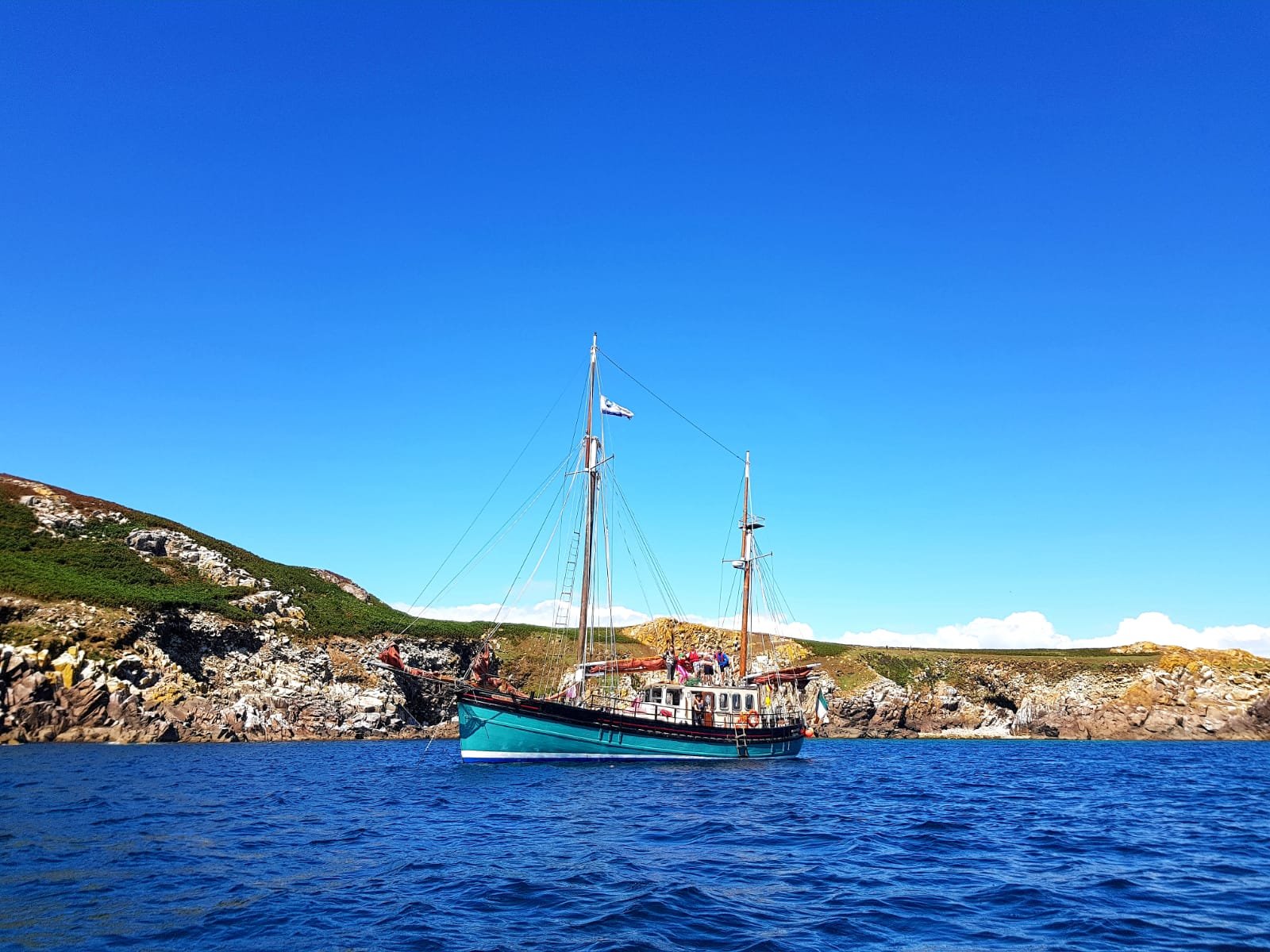 A sailing boat with two masts floating on calm blue water near rocky coastline with green and brown hills under a clear blue sky.