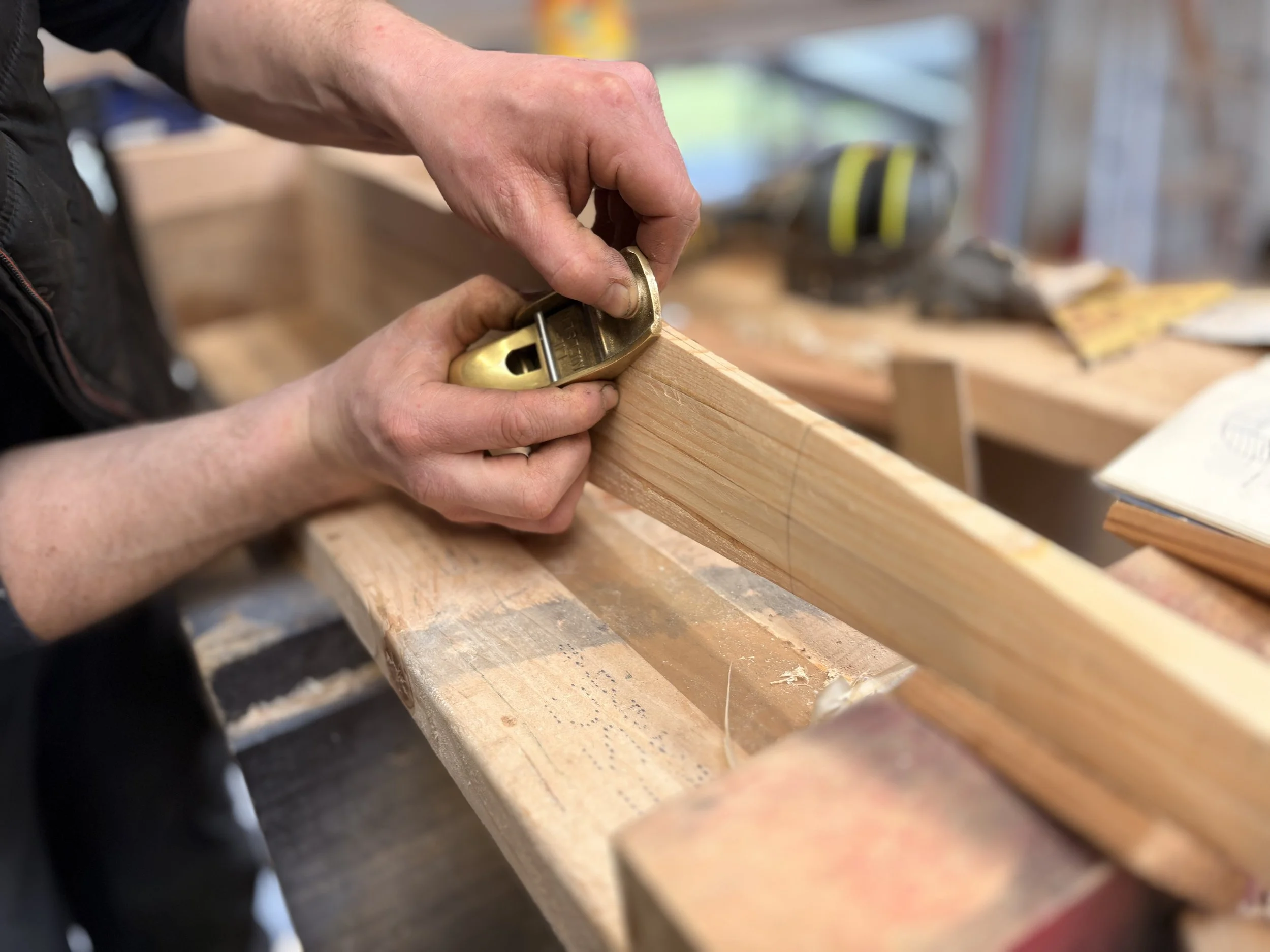 Close-up of hands using a hand planer to smooth a wooden board in a woodworking shop.