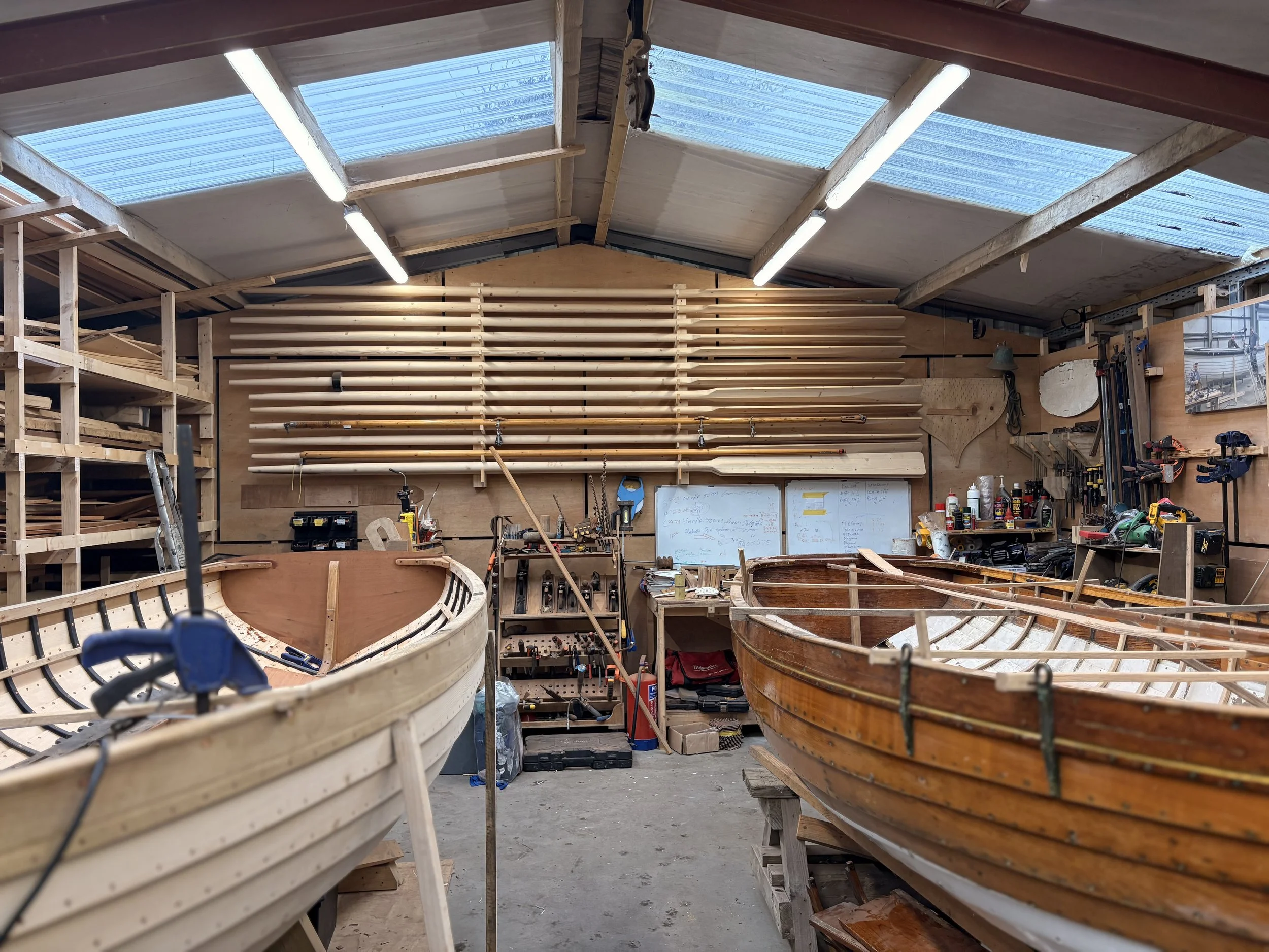 Interior of a woodworking shop with wooden boat frames under construction, tools and supplies on workbenches, wooden slats on the wall, and a partly completed boat in the foreground.