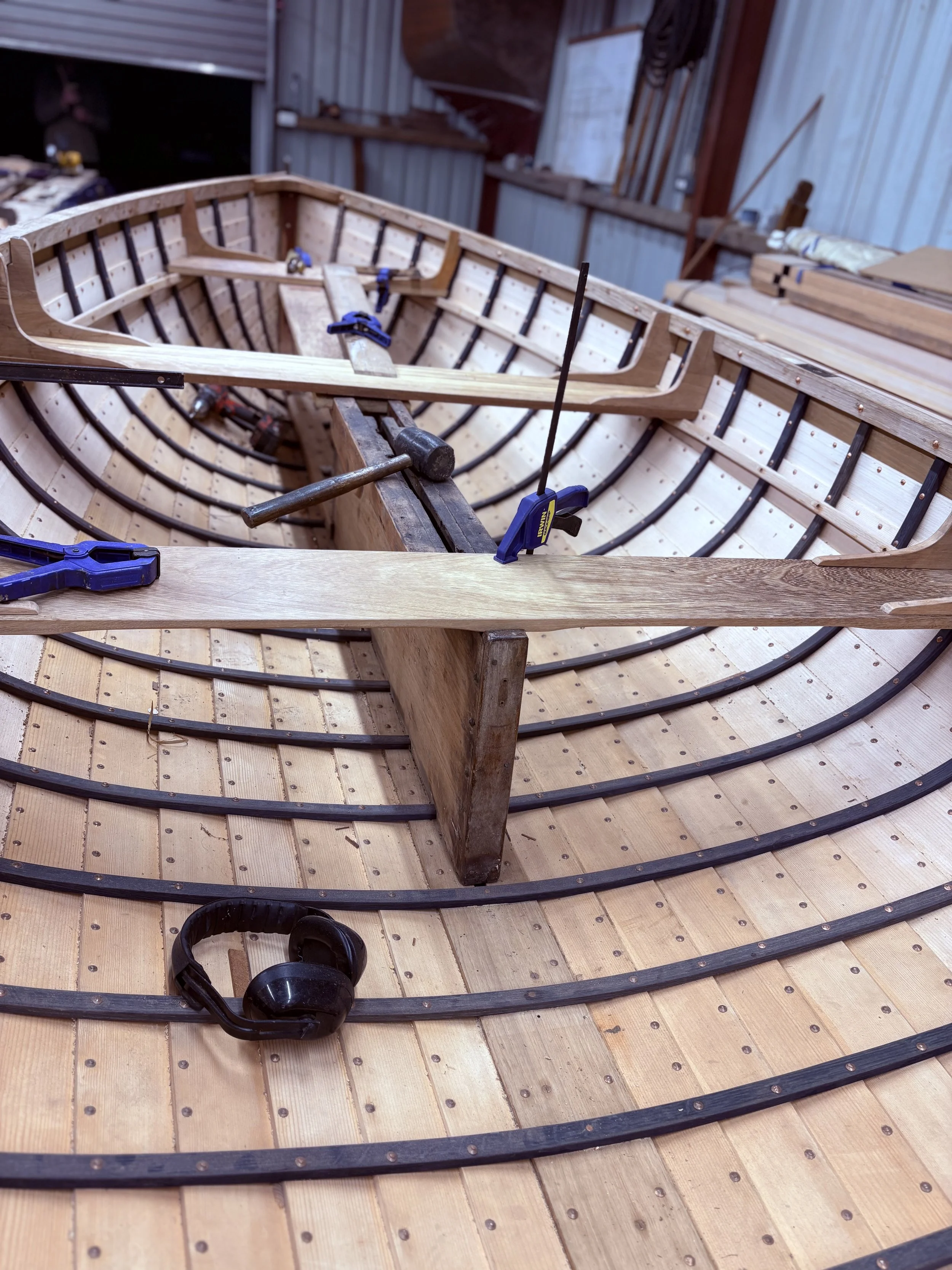 Under construction wooden boat in a workshop, with clamps and tools, and headphones resting on the wooden planks.