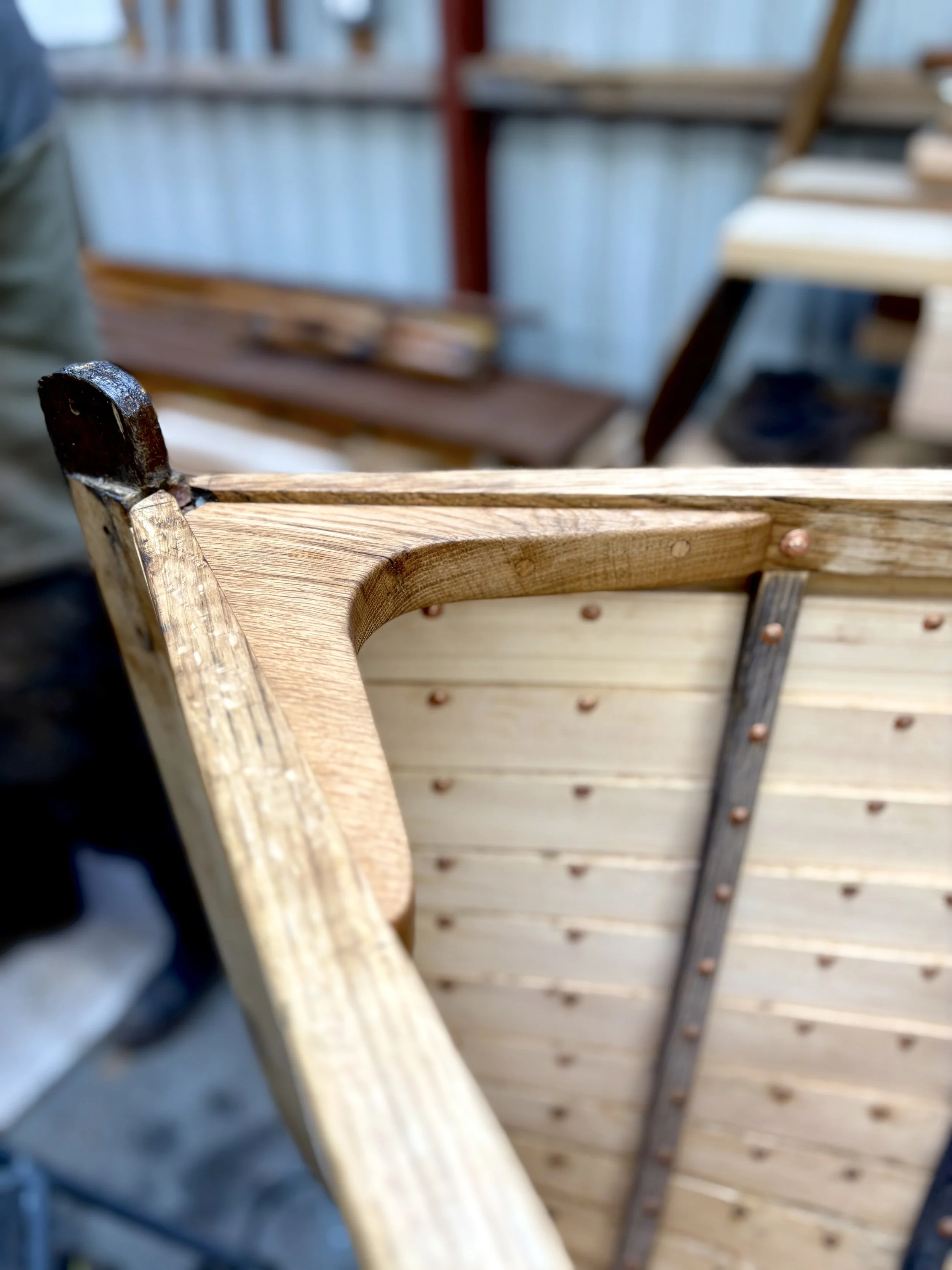 Close-up of a wooden boat under construction, showing the light-colored wooden planks and the dark metal nails securing them, with an out-of-focus workshop background.