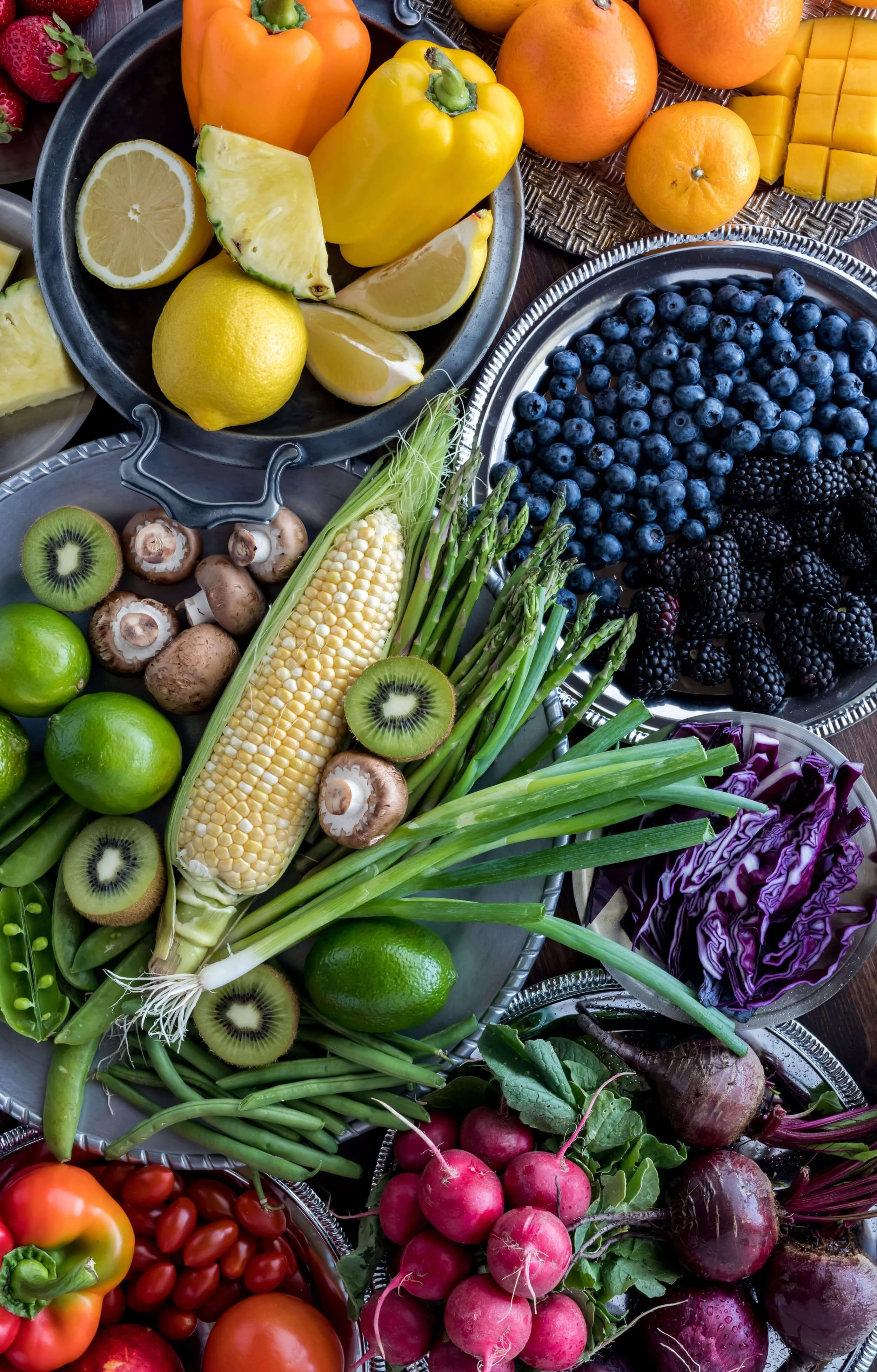 A colorful assortment of fresh fruits and vegetables, including bell peppers, lemons, oranges, strawberries, blueberries, blackberries, kiwis, corn, asparagus, radishes, cherry tomatoes, purple cabbage, and mushrooms displayed in metal and woven baskets.