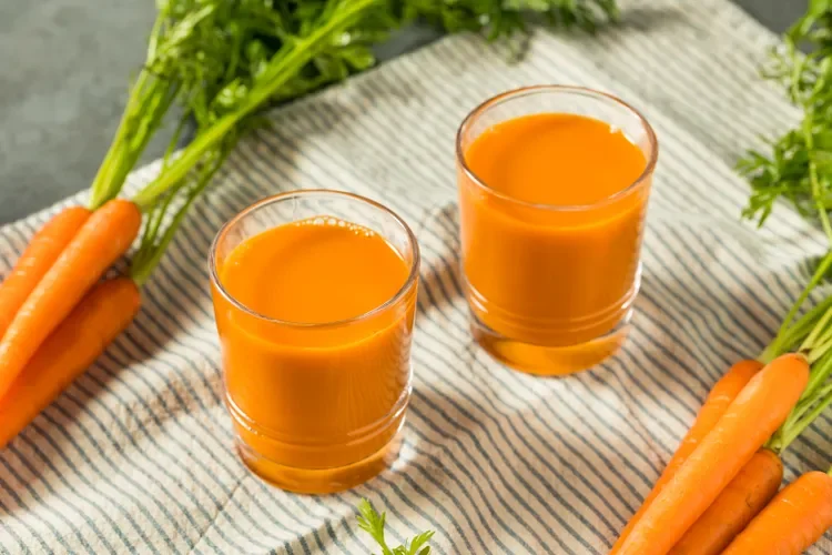 Two glasses of fresh carrot juice surrounded by whole carrots with green tops on a striped cloth.