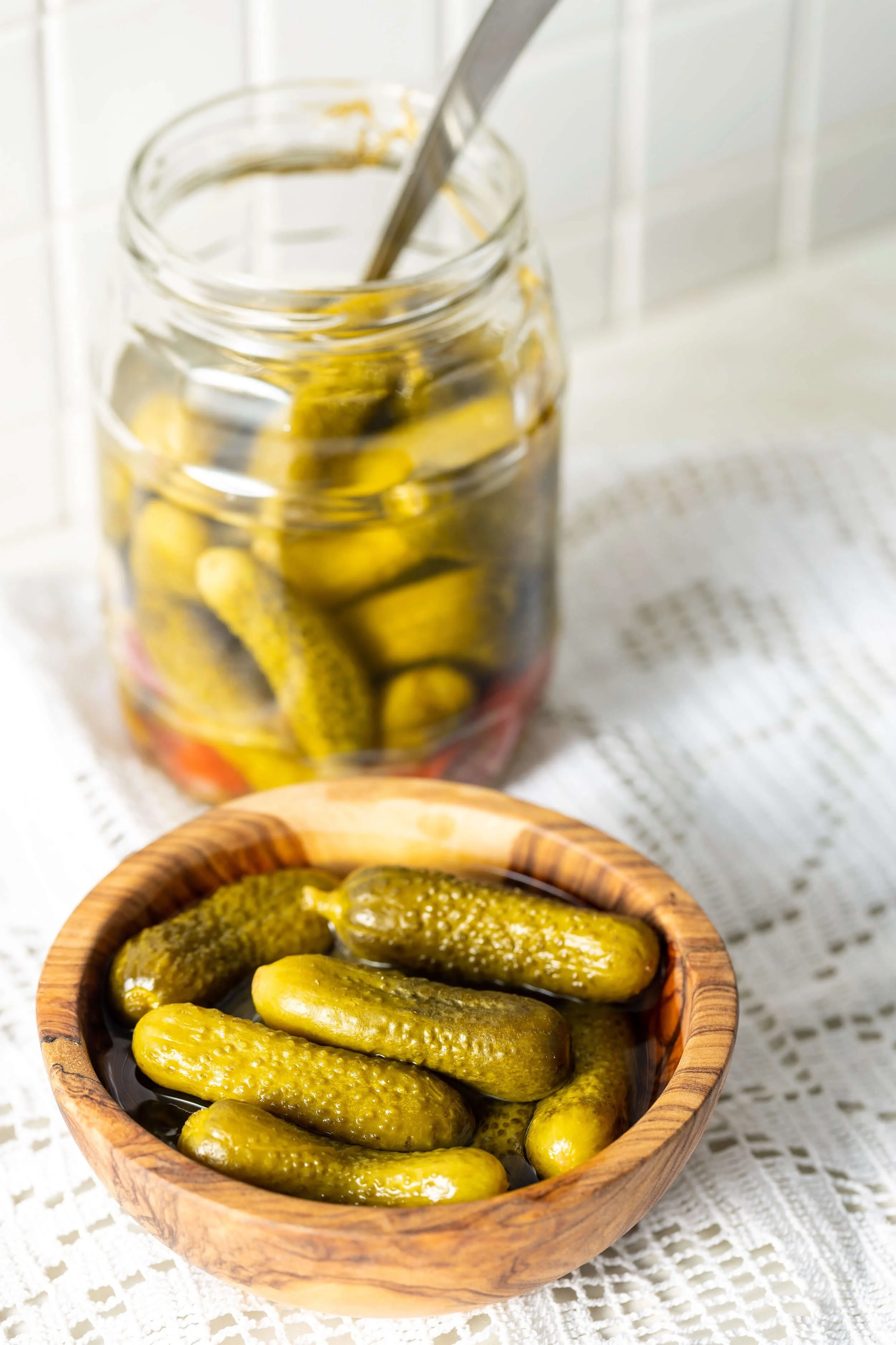 A wooden bowl of pickles and a glass jar filled with pickles on a white tablecloth.