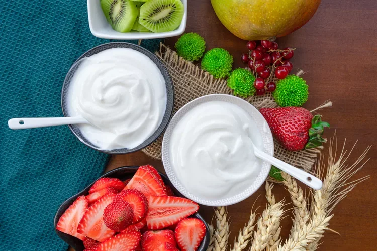 Bowl of yogurt, sliced strawberries, kiwi, apple, red grapes, green grapes, wheat stalks, and green decorative spheres on a wooden table.