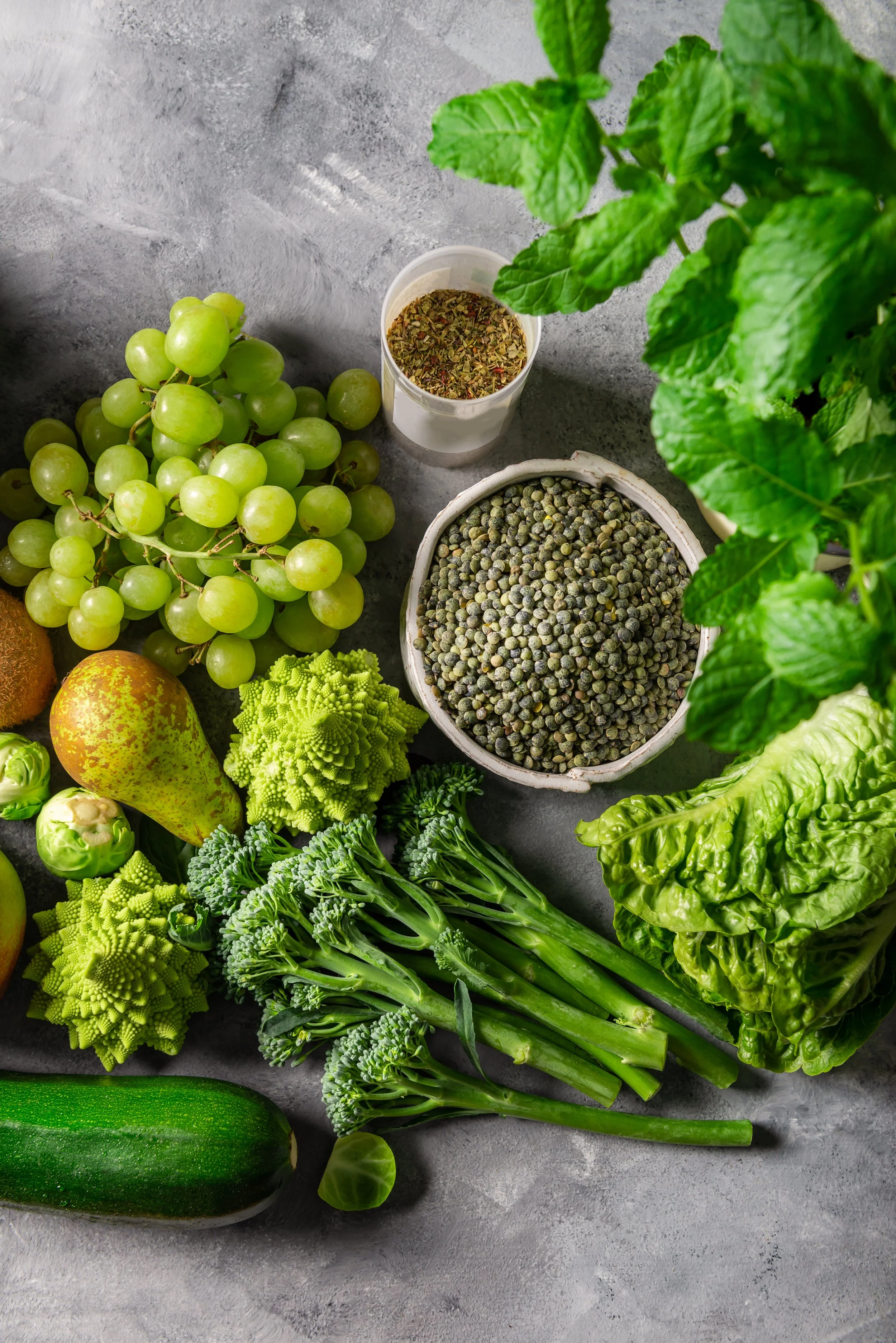 A variety of fresh green fruits, vegetables, and herbs on a gray surface, including green grapes, a cucumber, broccoli, lettuce, a pear, and small Brussels sprouts, along with bowls of herbs and spices.