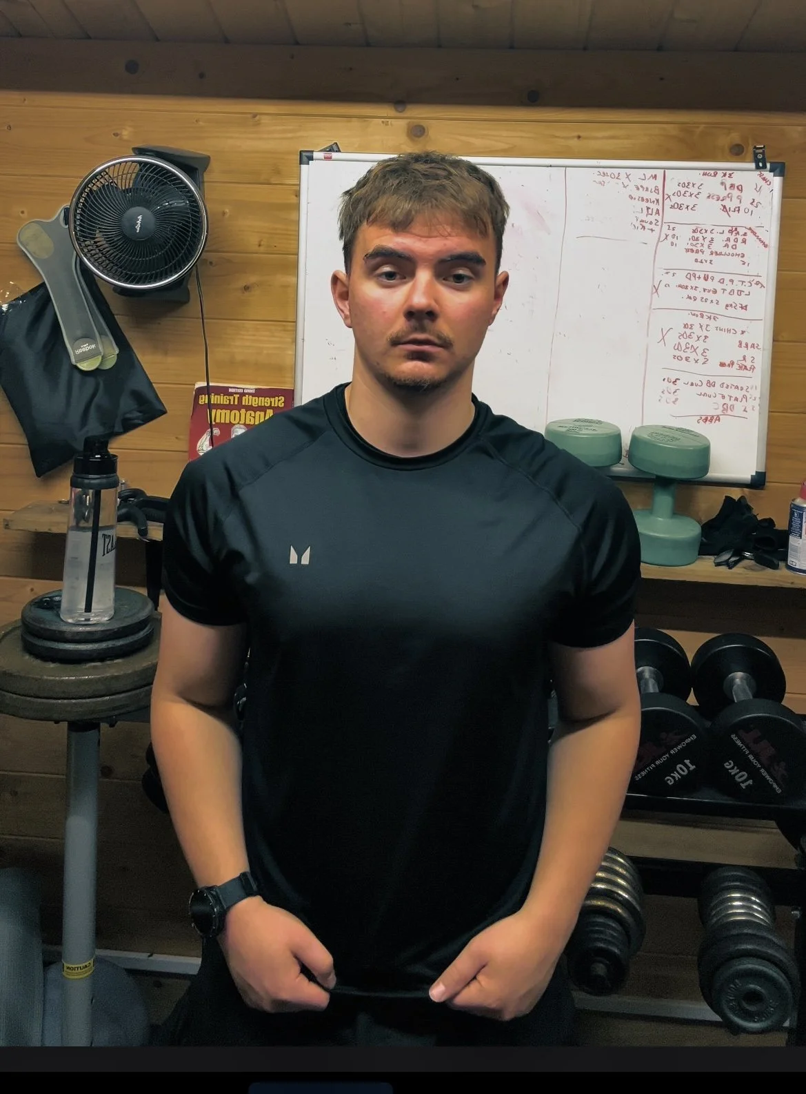 A young man in workout clothes standing in a gym with weightlifting equipment and a whiteboard in the background.