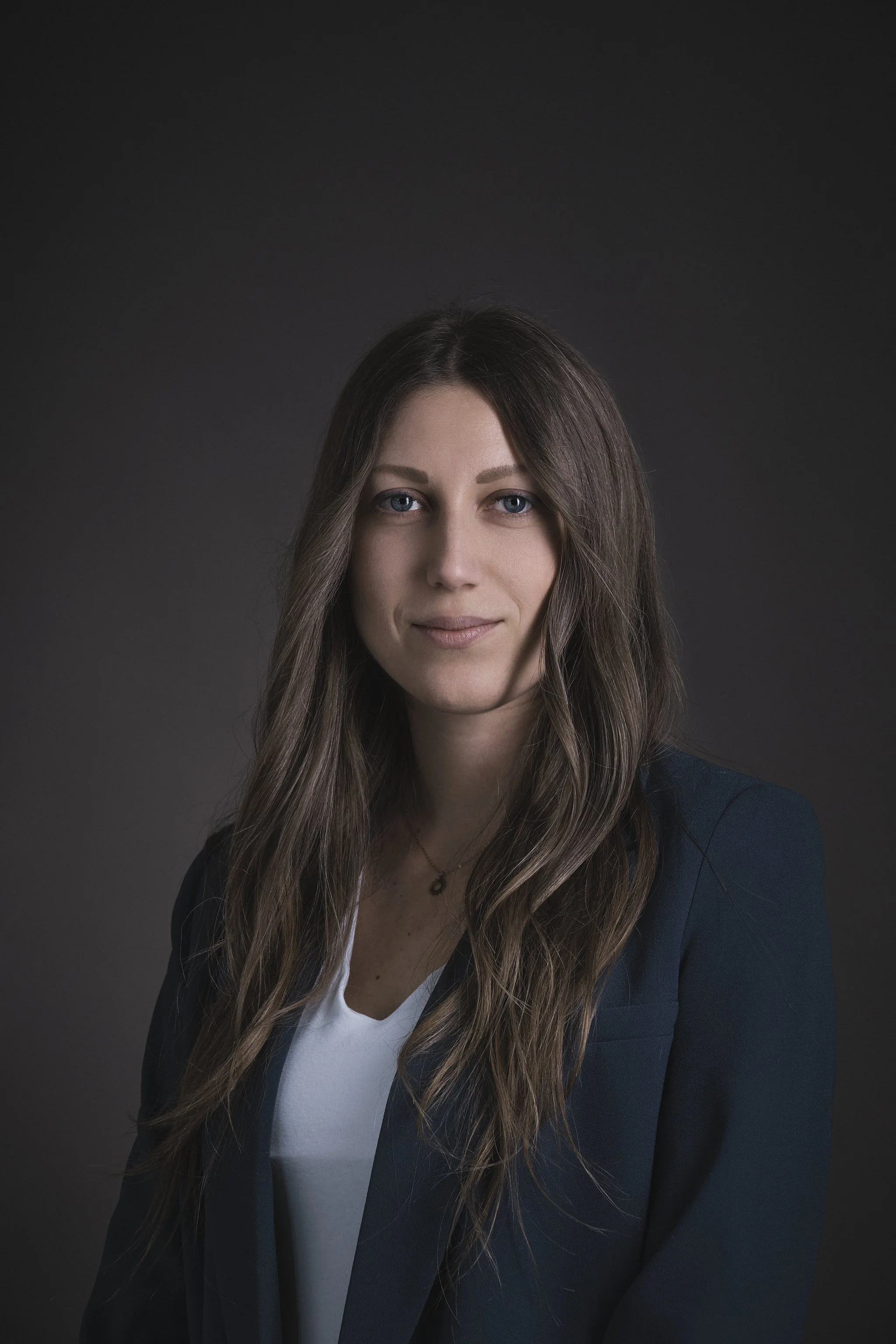 Una donna sorridente seduta su un divano, con capelli castani, indossa un maglione nero e pantaloni bianchi.