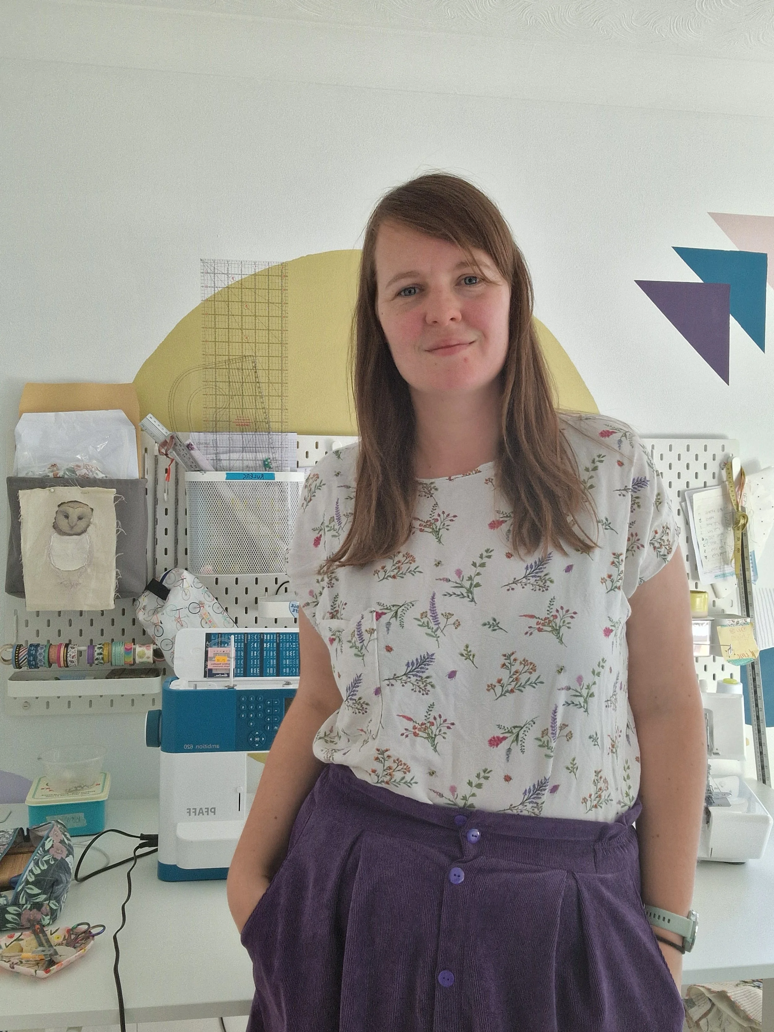 A woman stands in front of a desk looking at the camera. The desk has a sewing machine and tools on it.