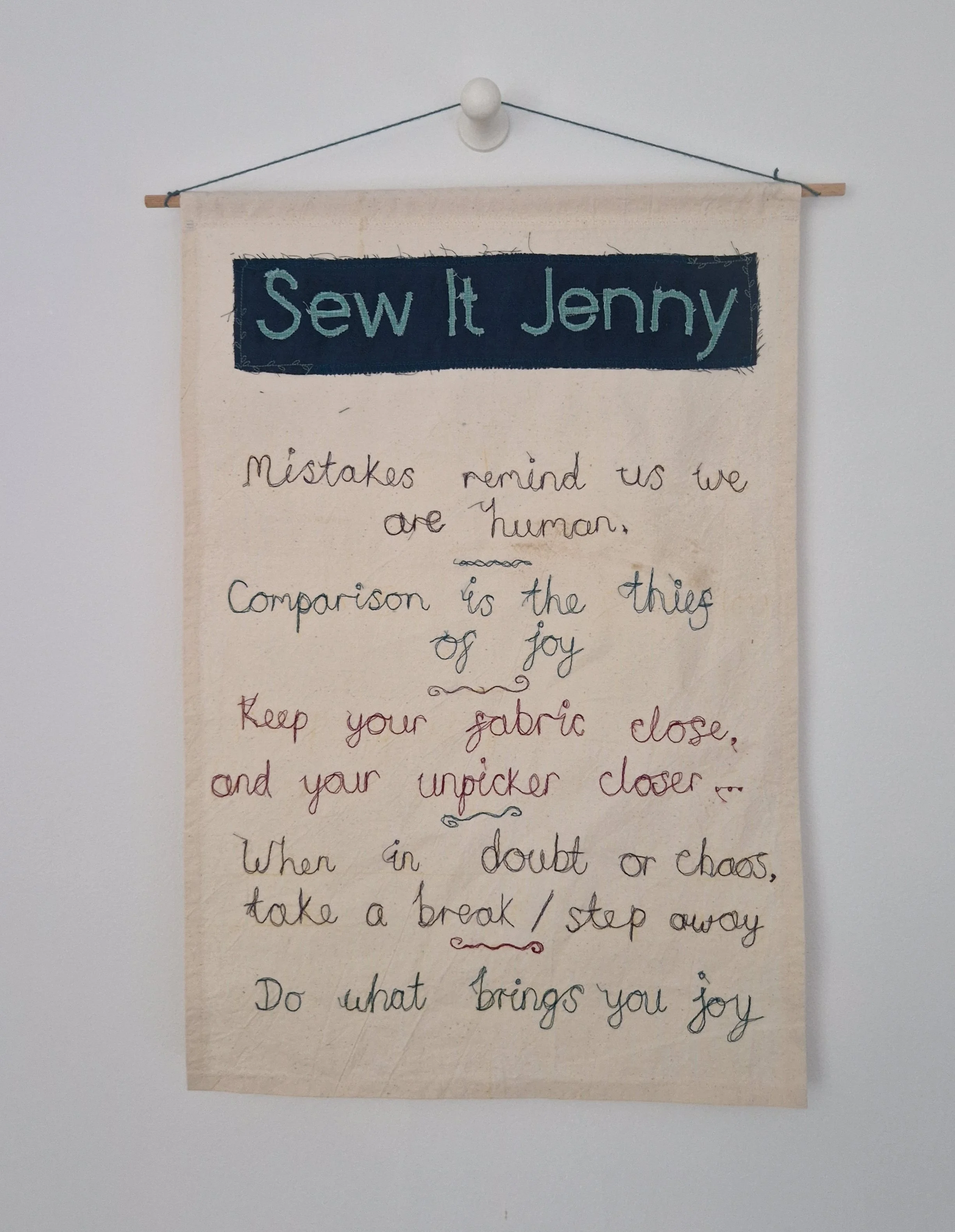 A fabric wall hanging with a dark blue rectangular patch at the top with the words 'Sew It Jenny' embroidered in light blue. Handwritten quotes about mistakes, joy, and taking breaks are written beneath the patch.