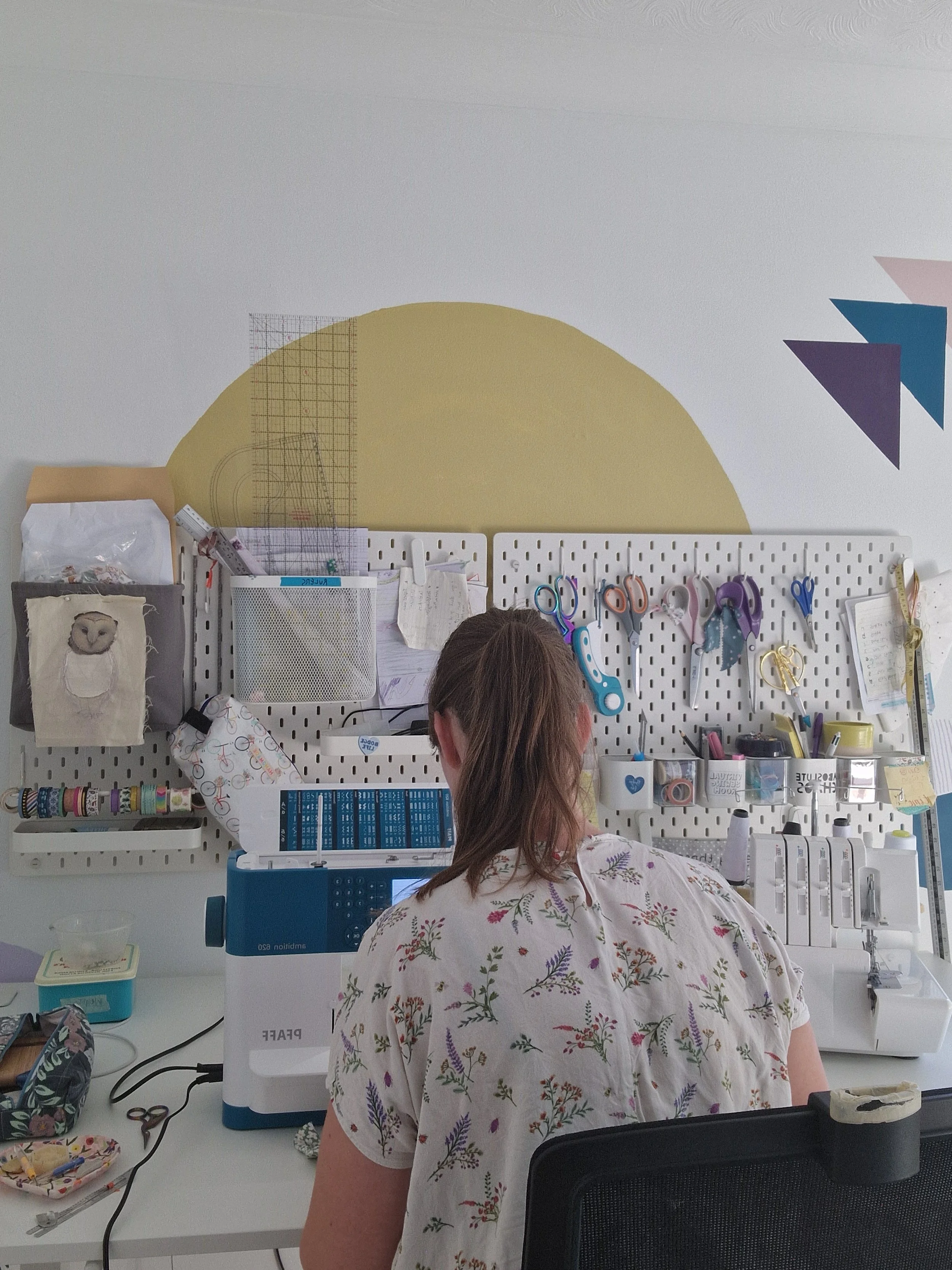 Person working at a sewing or embroidery machine in a craft room with various supplies and scissors on a pegboard behind them.