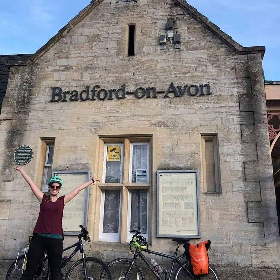 A woman wearing sunglasses and a green bike helmet stands in front of a stone building labeled 'Bradford-on-Avon' with her arms raised. There are two bicycles parked against the wall, one has an orange bag. The building has several signs, including a CCTV warning and other informational plaques.