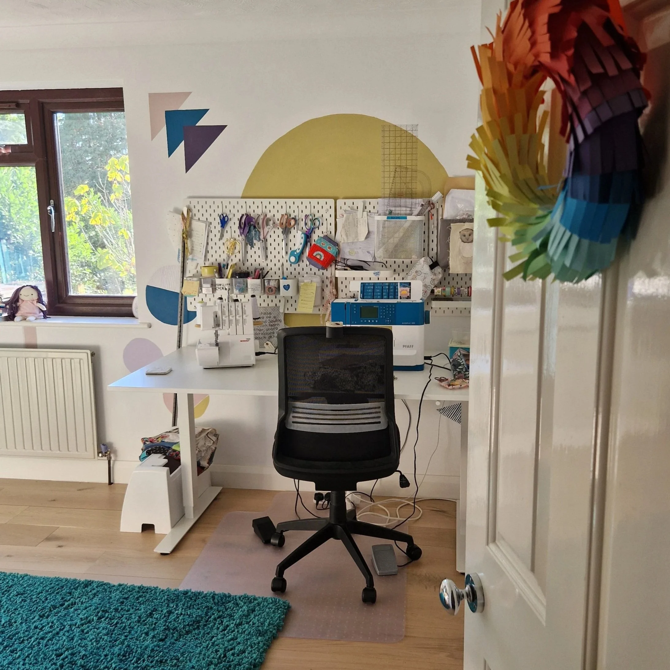 Home sewing or craft room with a white desk, black swivel chair, sewing machine, and pattern paper on a pegboard wall. Rainbow paper chain decoration on the door frame, a blue rug, and a window with greenery outside.