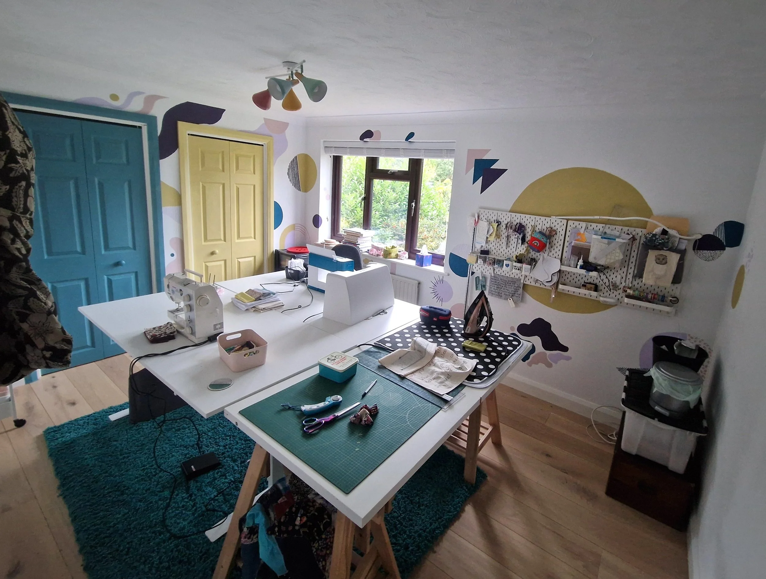 A craft room with two work tables, sewing materials, and colorful wall decorations featuring abstract shapes and a large yellow semi-circle. There are blue and yellow closet doors and a window with greenery outside.
