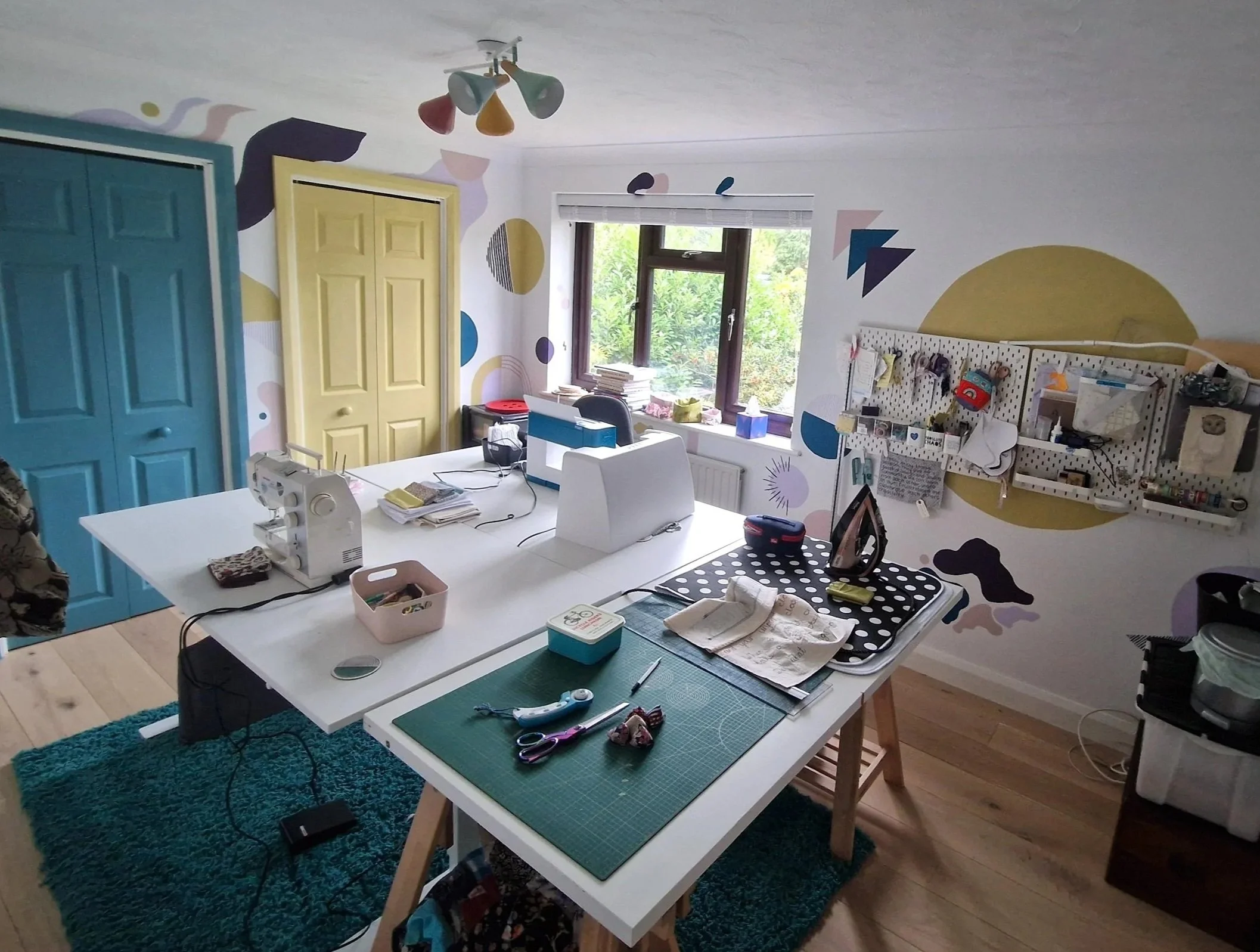 A craft room with a white central table covered in sewing supplies, scissors, and fabric. The wall features colorful geometric and abstract painted patterns. There are two closets, one painted turquoise and another yellow, and a window with greenery outside. Pegboards on the right wall hold various tools and materials.