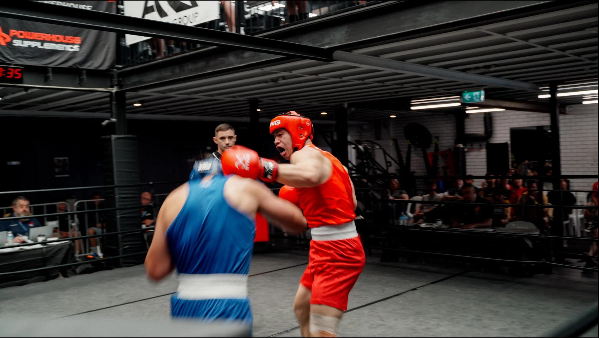 Boxers sparring in a ring during a match, with a referee observing, in a gym or sports venue filled with spectators.
