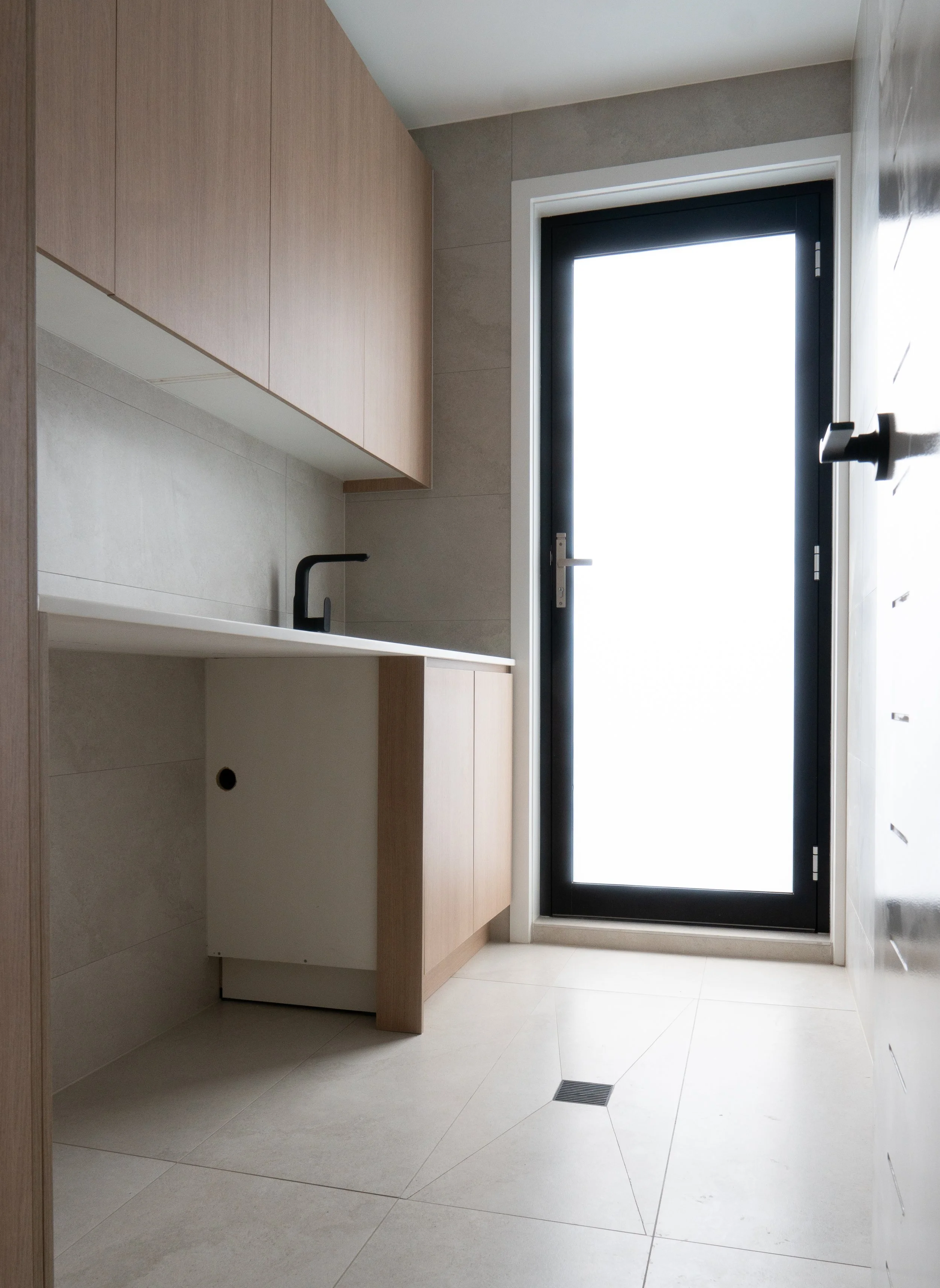 Empty laundry room with beige tiles, wooden cabinets, black sink faucet, and a large glass door leading outside