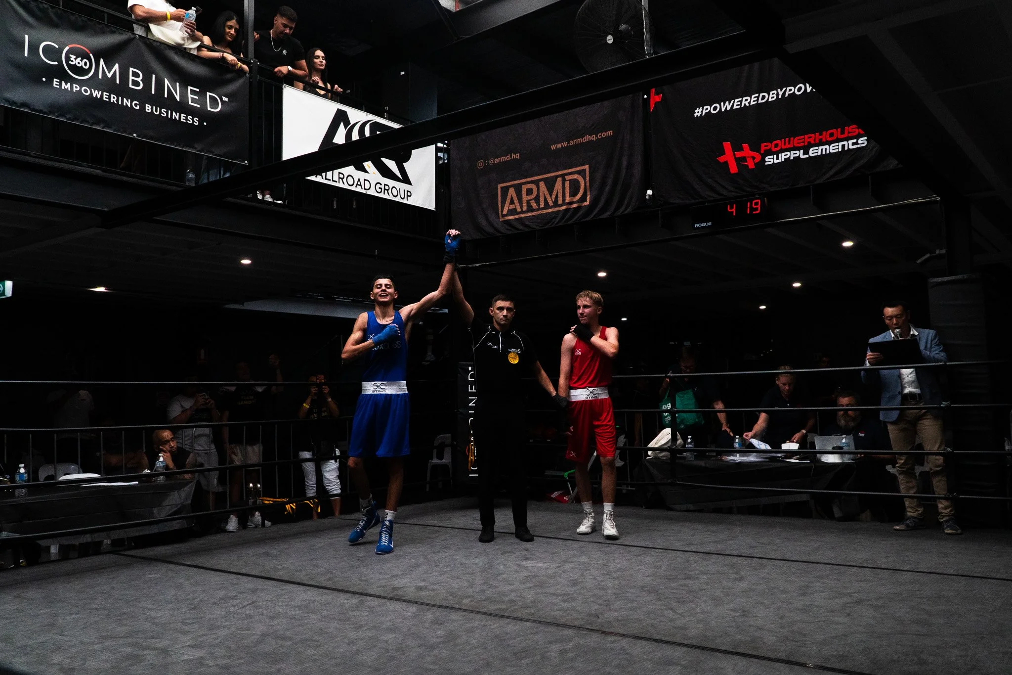 Two male boxers, one in blue and one in red, standing in a boxing ring after a match. The boxer in blue is smiling and raising his arm in victory, while the boxer in red looks down. The referee is in the middle, holding the winner's hand. Spectators 