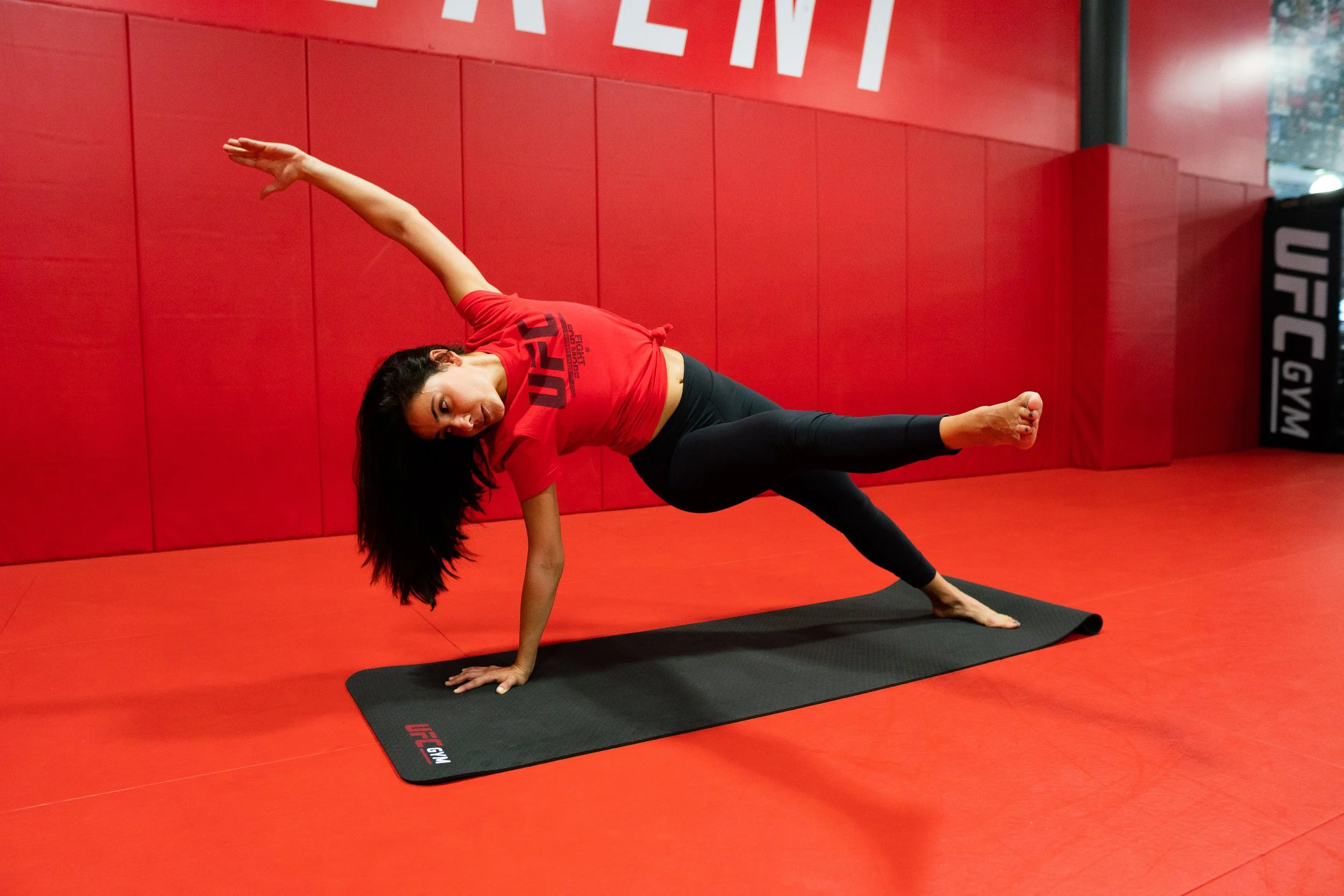 A woman practicing yoga on a black mat at a UFC gym, performing a side plank variation with one hand on the mat and the other arm extended overhead, in a red gym with UFC logos.