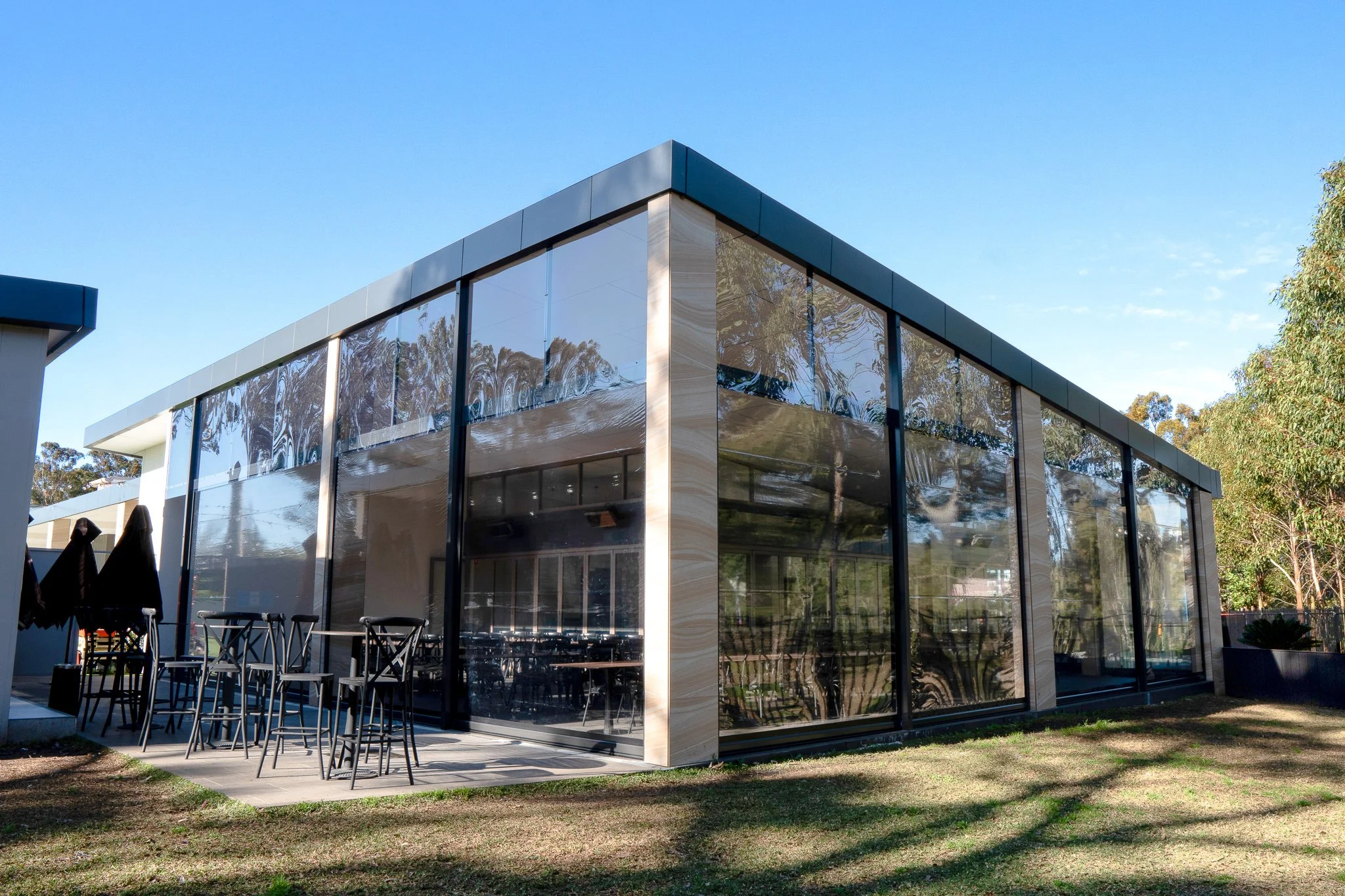 Modern glass building with patio seating and outdoor umbrellas, surrounded by trees and a clear blue sky.