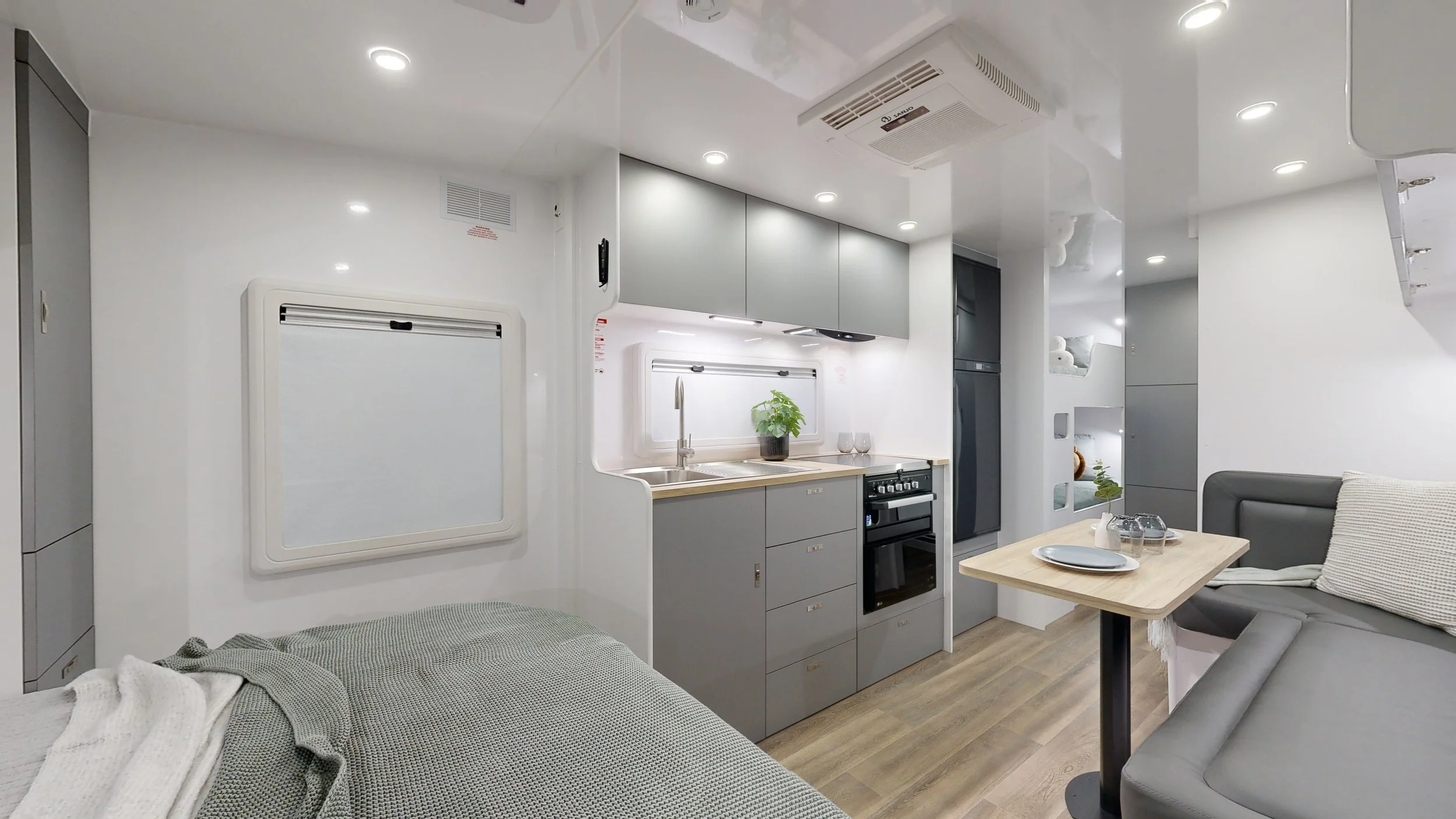 Compact living space featuring a small kitchen with gray cabinets, a sink, a stovetop, and a potted plant. Adjacent is a dining area with a table set with a plate, glass, and white napkin. A gray L-shaped sofa with a pillow is nearby, and a bed is pa