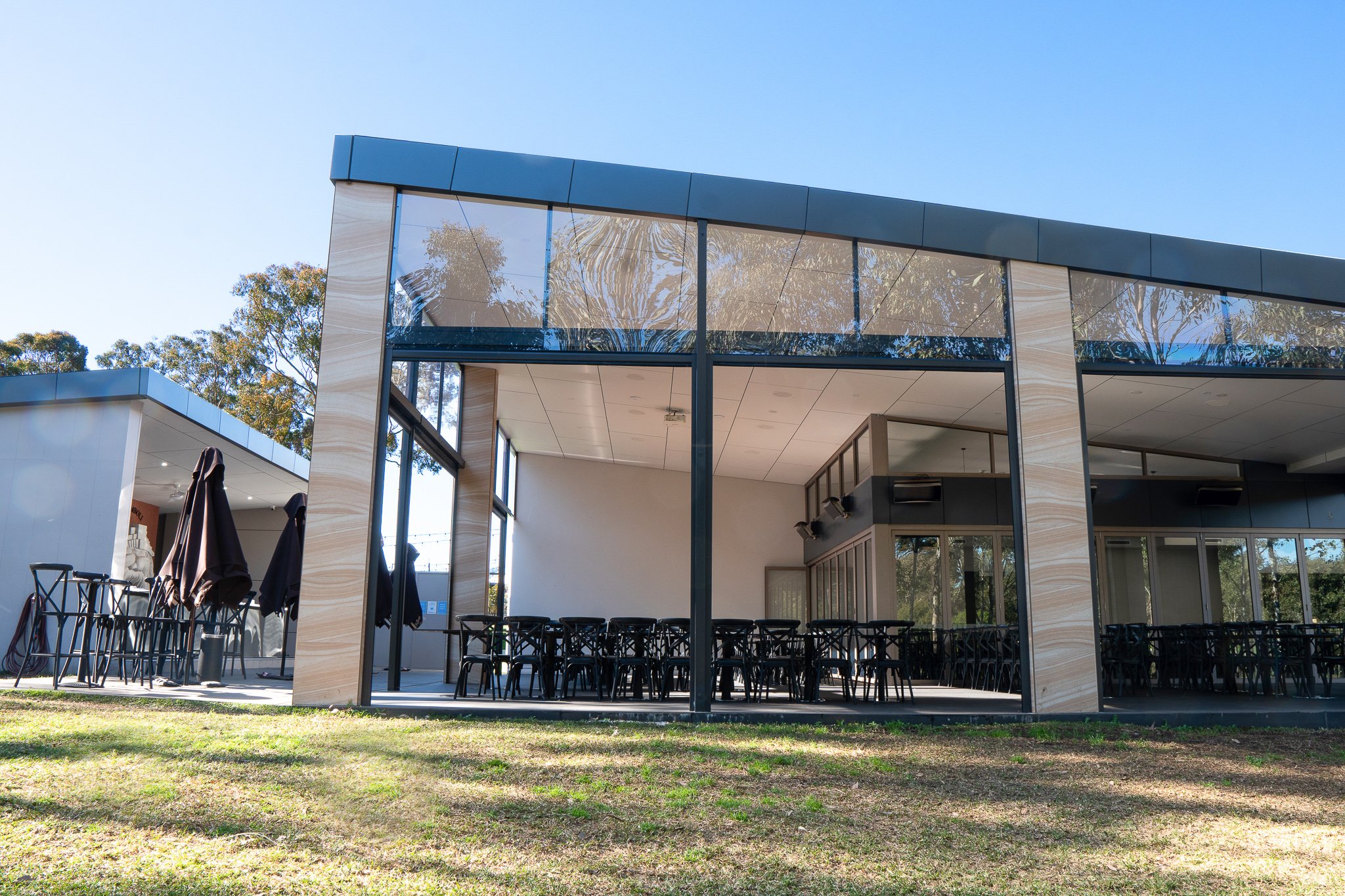 Modern building with glass windows, outdoor seating with black chairs, and umbrellas, surrounded by trees and grass