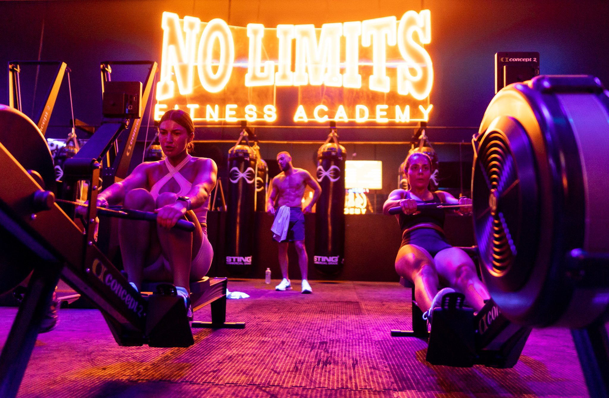 Inside a dimly lit fitness studio with neon orange sign that reads 'NO LIMITS FITNESS ACADEMY'. Two women are using rowing machines, and a man, holding a towel, stands in the background near punching bags.