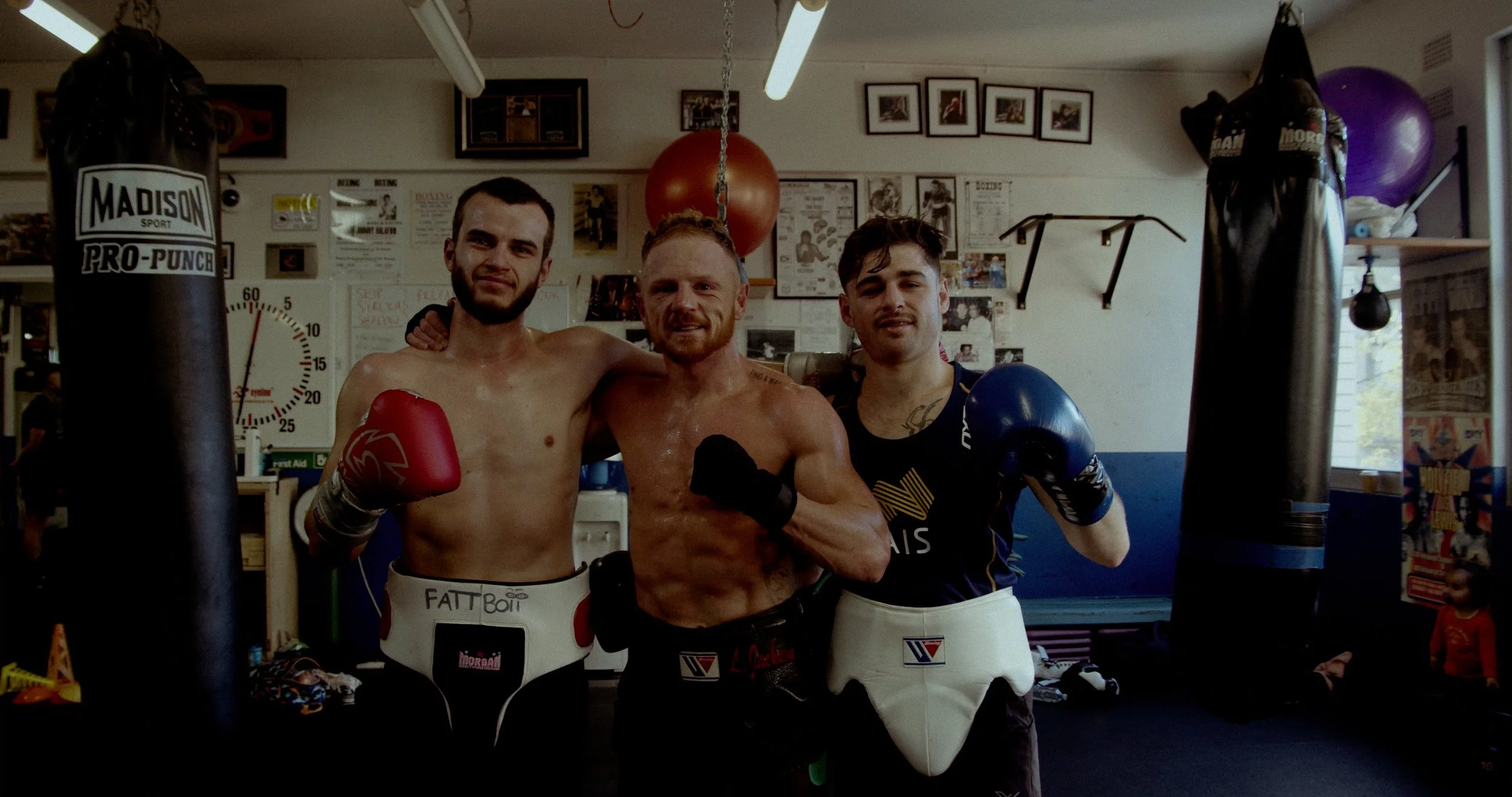 Three male boxers pose inside a boxing gym, smiling and showing their boxing gloves, with punching bags or training equipment behind them.