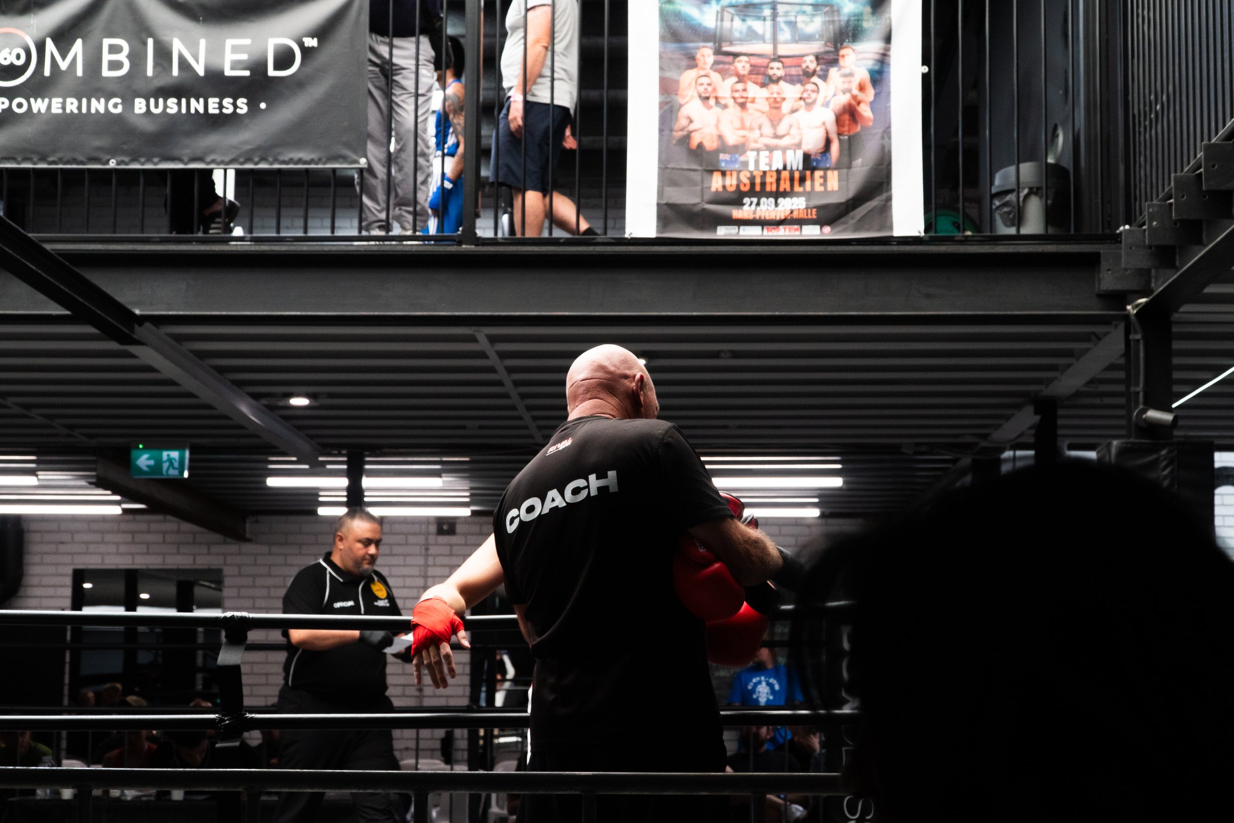 Boxing gym with a coach and a boxer training in the ring, and spectators watching from the upper level, promotional posters on the wall.