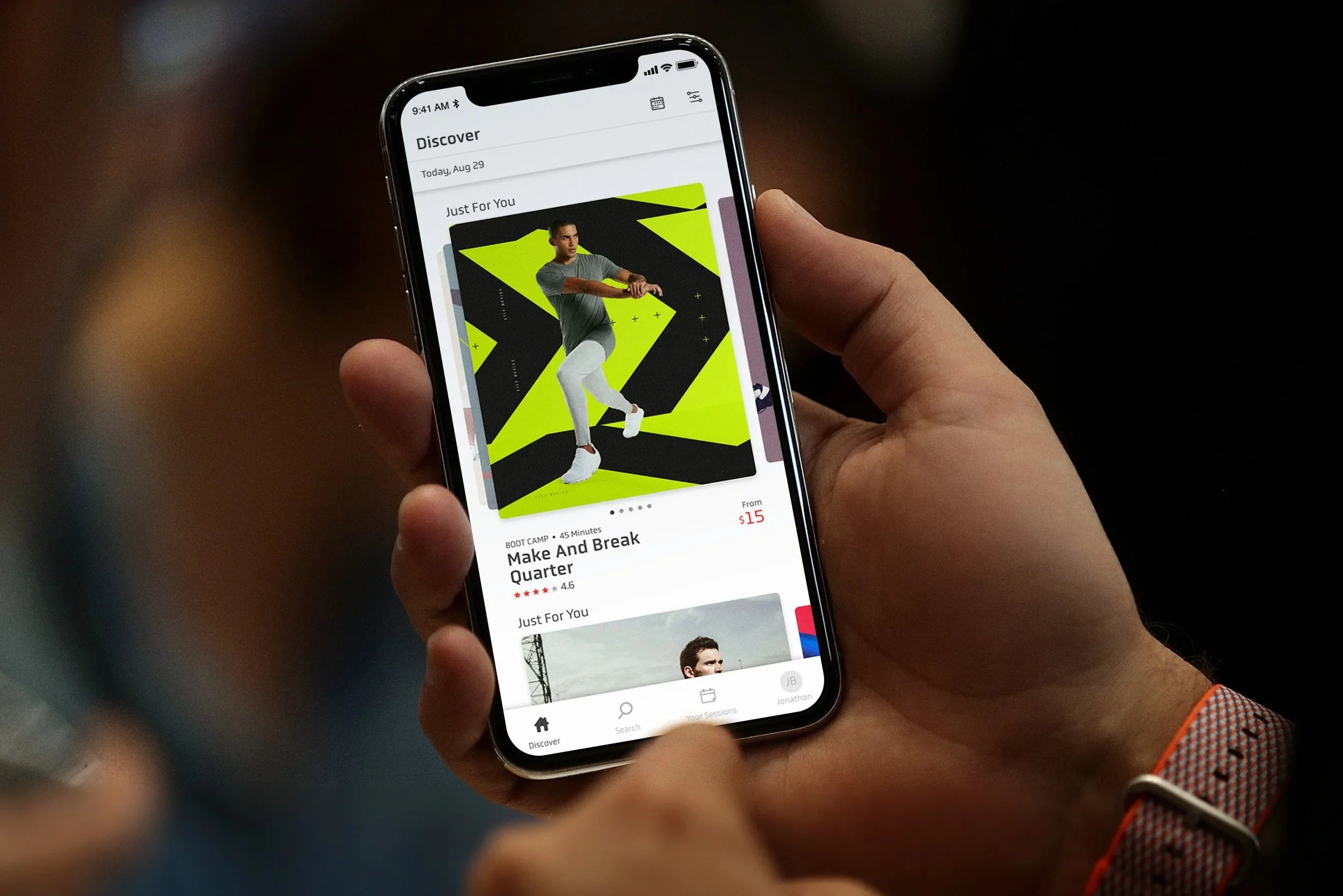 Person holding a smartphone displaying a fitness class app with a man in workout attire on the screen.