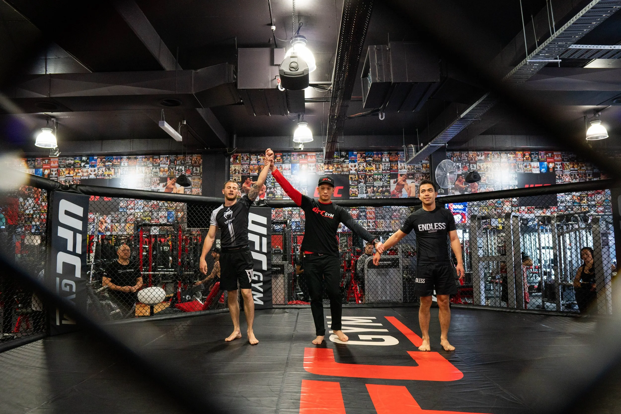 Two fighters in MMA uniforms with a referee in the center raising the winner's hand in an MMA gym.