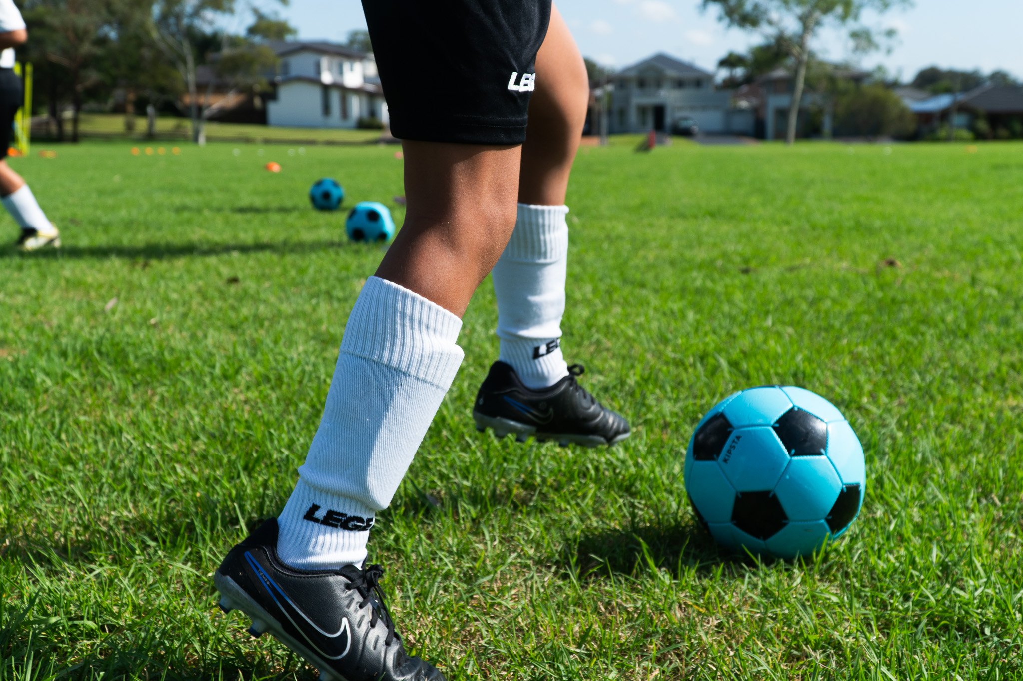 Child in black soccer shorts, white socks, and black Nike cleats practicing soccer on a green field with a blue and black ball nearby, other kids in the background.