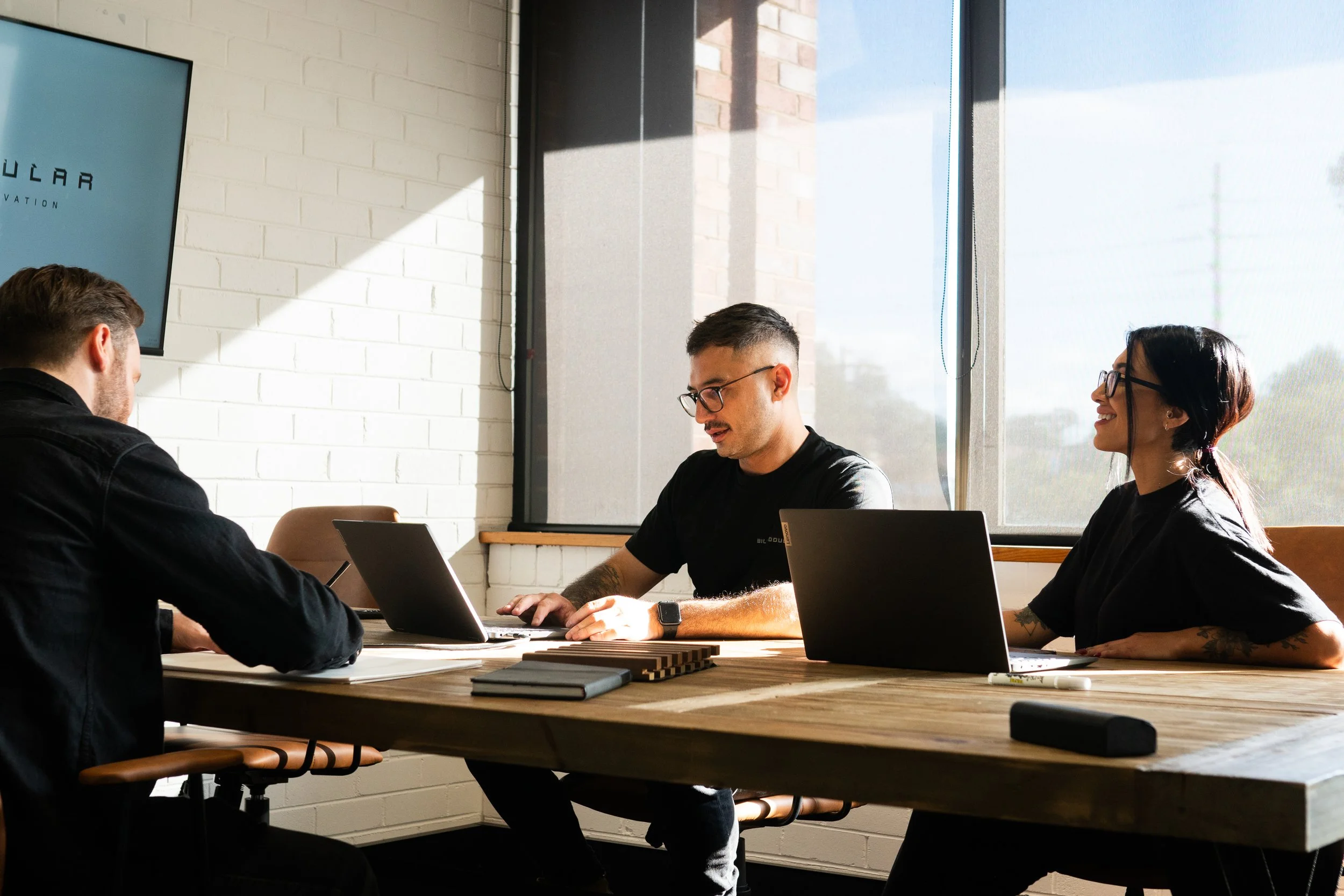 Three people sitting at a wooden table in a modern, well-lit office room with large windows, working on laptops and having a discussion.