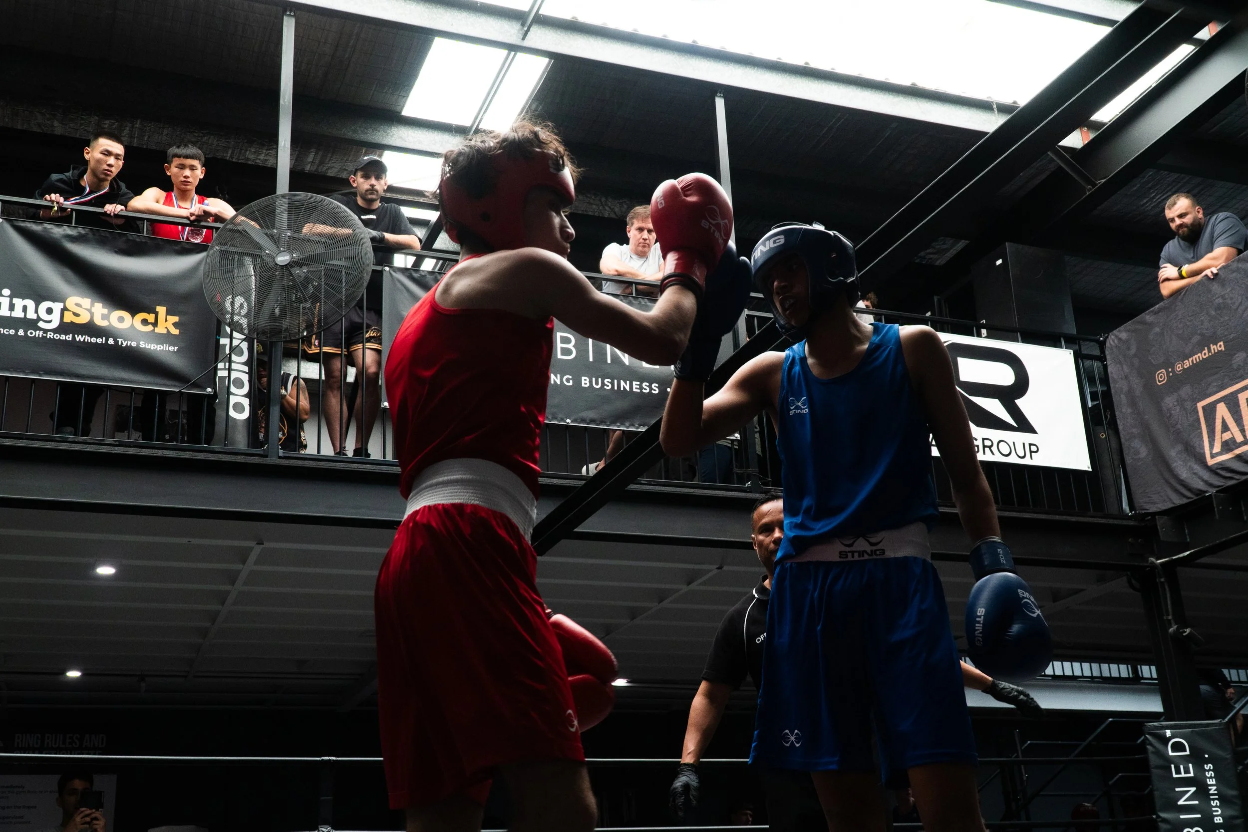 Two amateur boxers in red and blue shorts, wearing protective headgear and gloves, engaged in a match inside a boxing ring with spectators watching from the upper level.