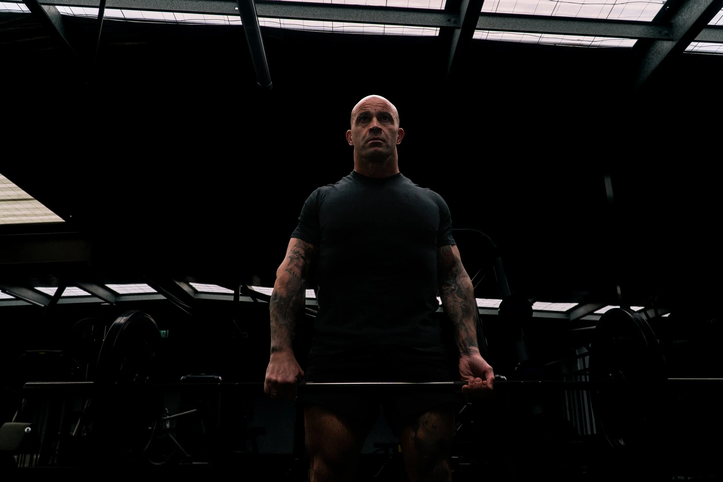 A muscular man with tattoos on his arms is lifting a barbell in a dark gym, with only the ceiling lights illuminating him.
