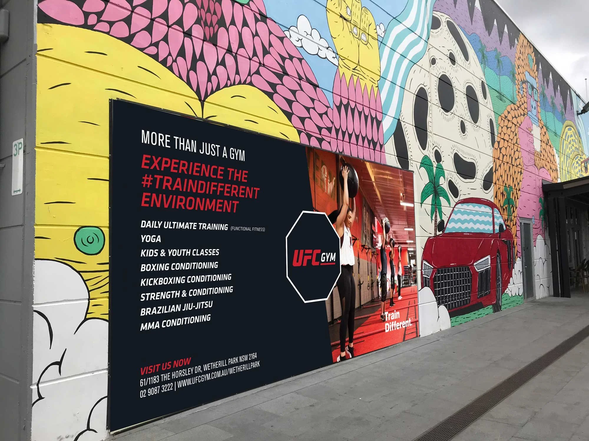 A colorful mural on the wall of a gym building with various abstract designs including a car, a skull, fruits, and animals. In front, a digital sign promotes UFC Gym services, featuring a woman exercising with a medicine ball and text detailing classes and contact information.