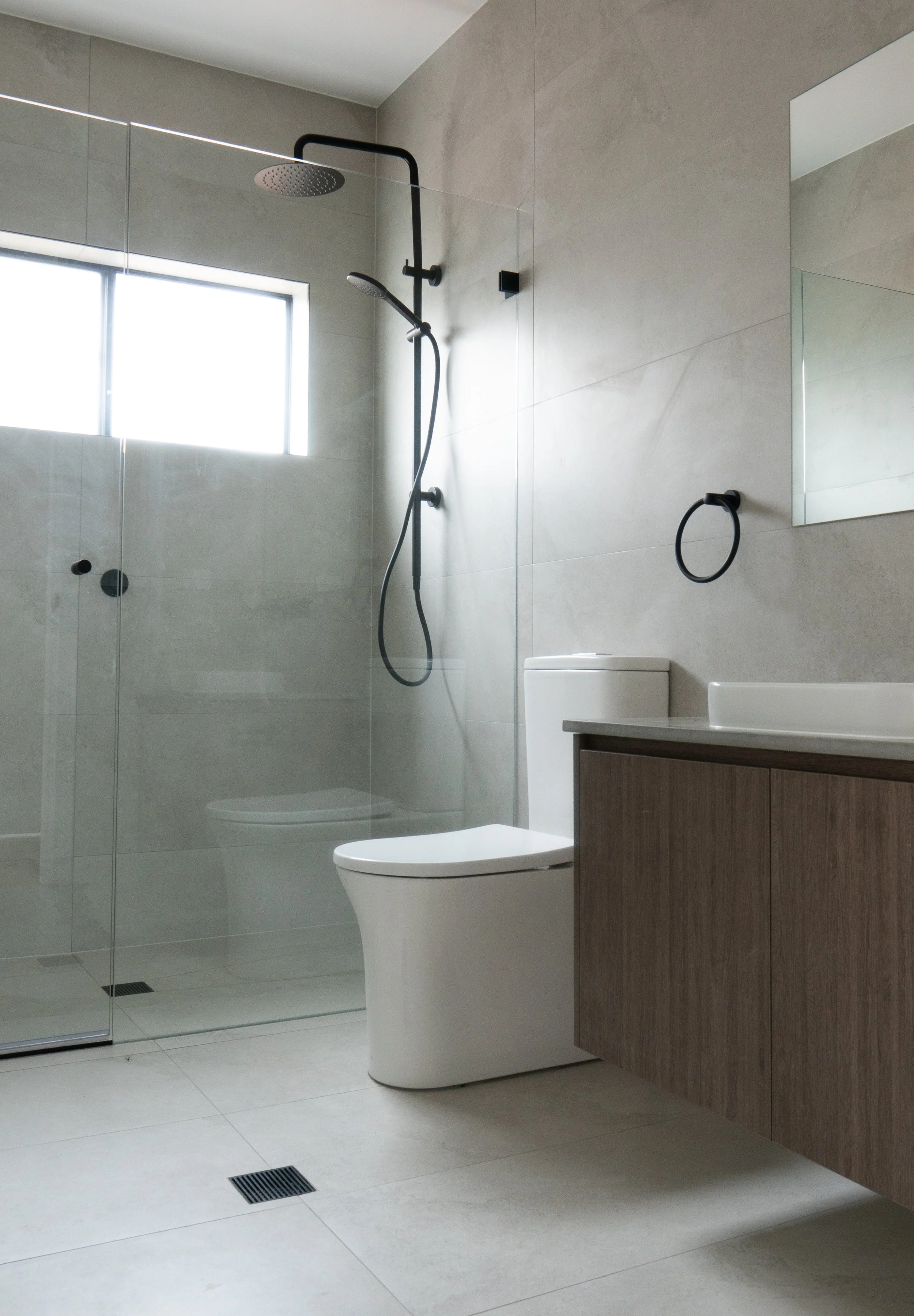 Modern bathroom with a walk-in shower, a white toilet, a wooden vanity with a sink, a mirror, and a towel ring, illuminated by natural light from a frosted window.