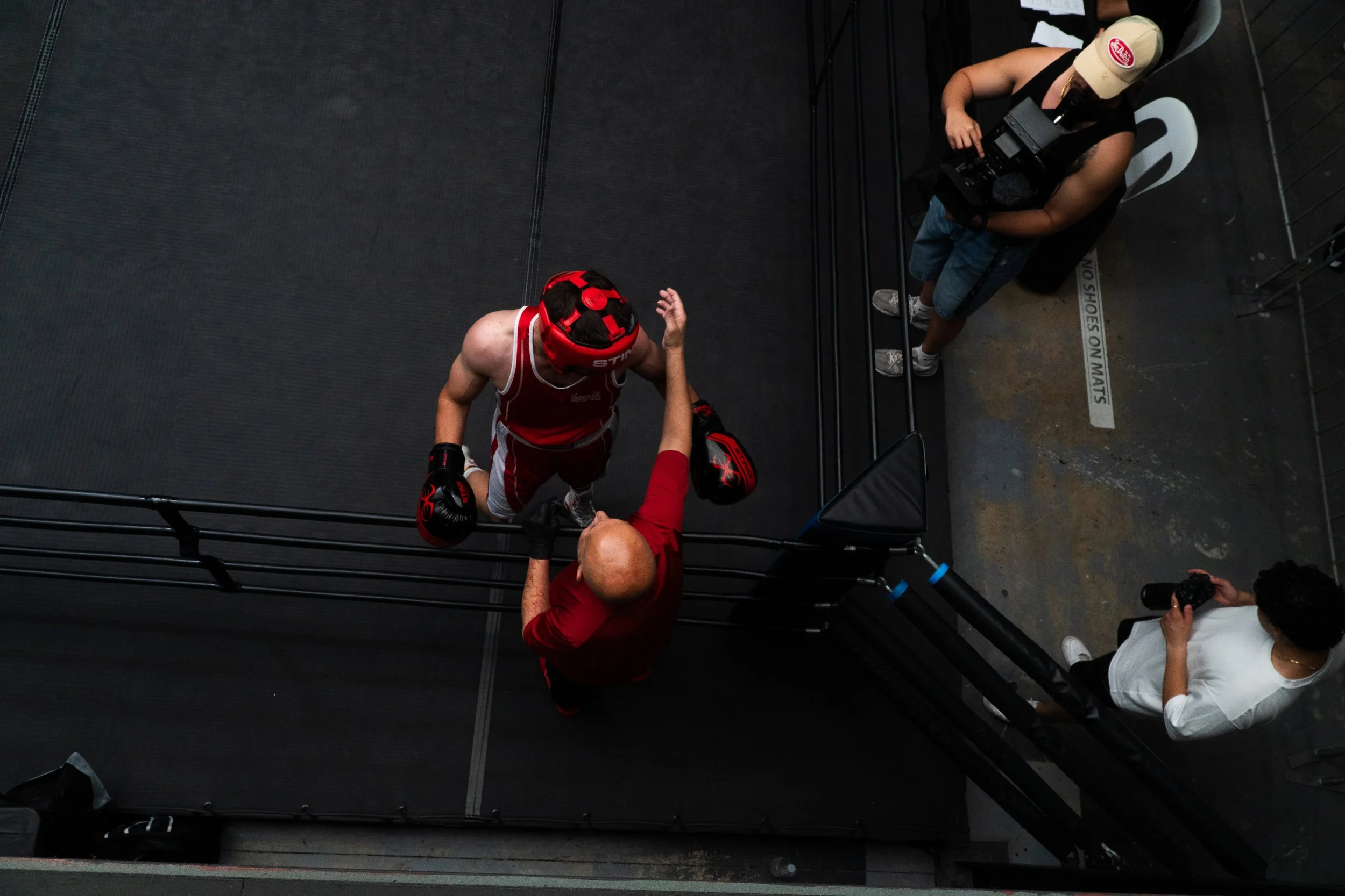 A boxer in red attire and boxing gloves is shaking hands with a bald man in red shirt, inside a boxing ring. Two individuals are observing; one is taking a photograph, and the other is standing near the ring.