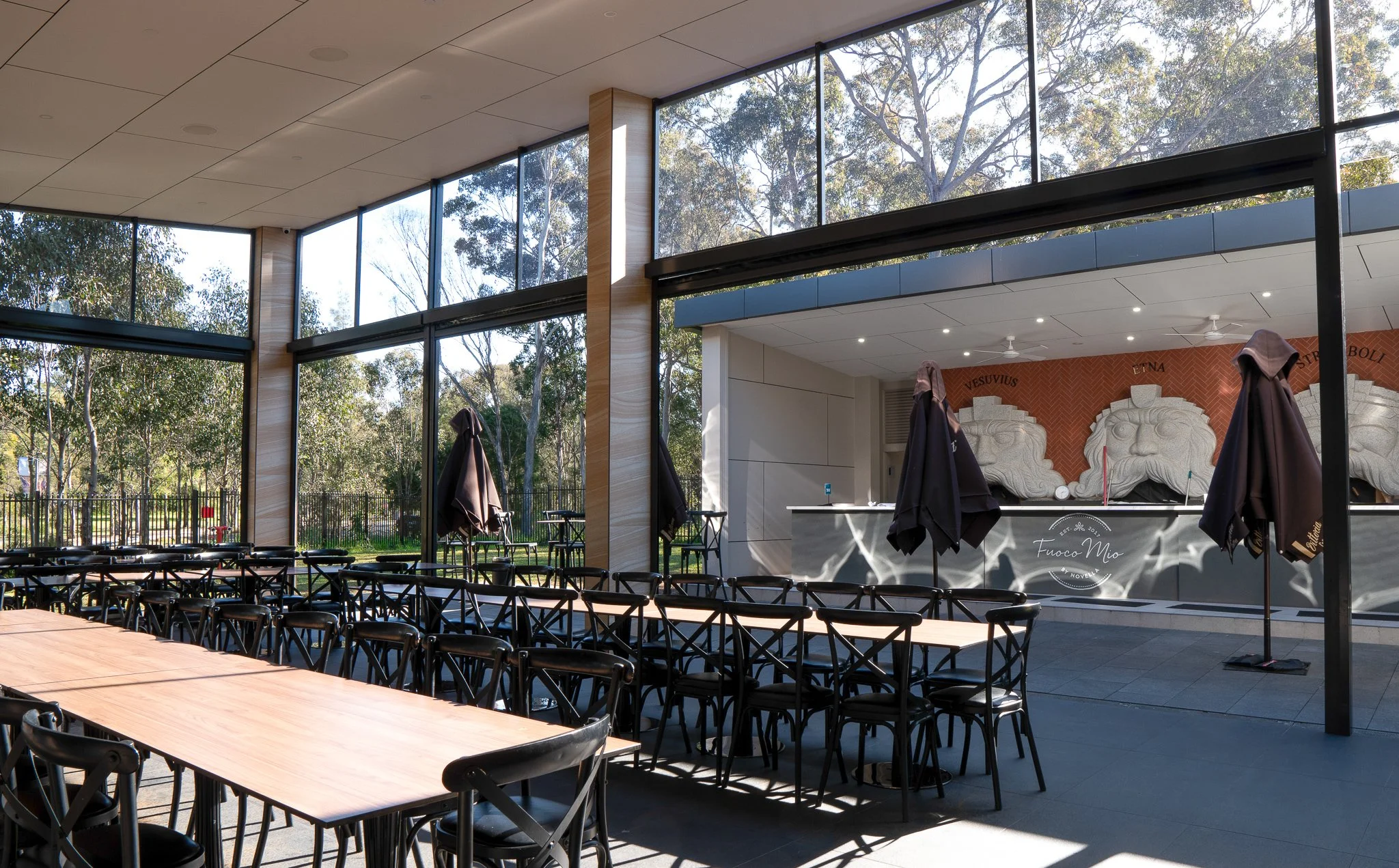 Interior of a modern restaurant or cafe with large glass windows, black chairs, wooden tables, and a counter area featuring carved sculptures and a sign that says 'Fuoco Mio'.