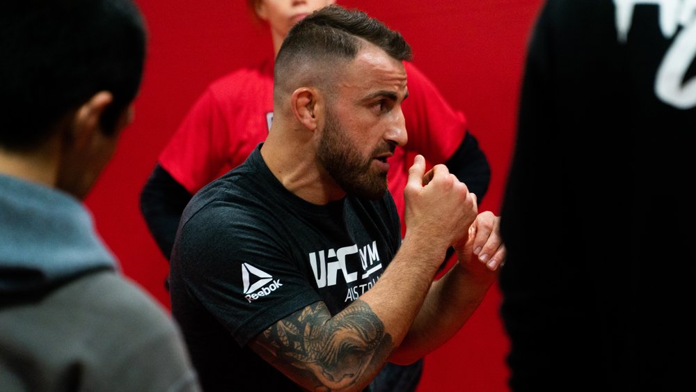 A man with a beard and tattoos on his left arm, wearing a black UFC T-shirt, is in a boxing stance with his fists raised, inside a gym with red walls. Other people are around him.