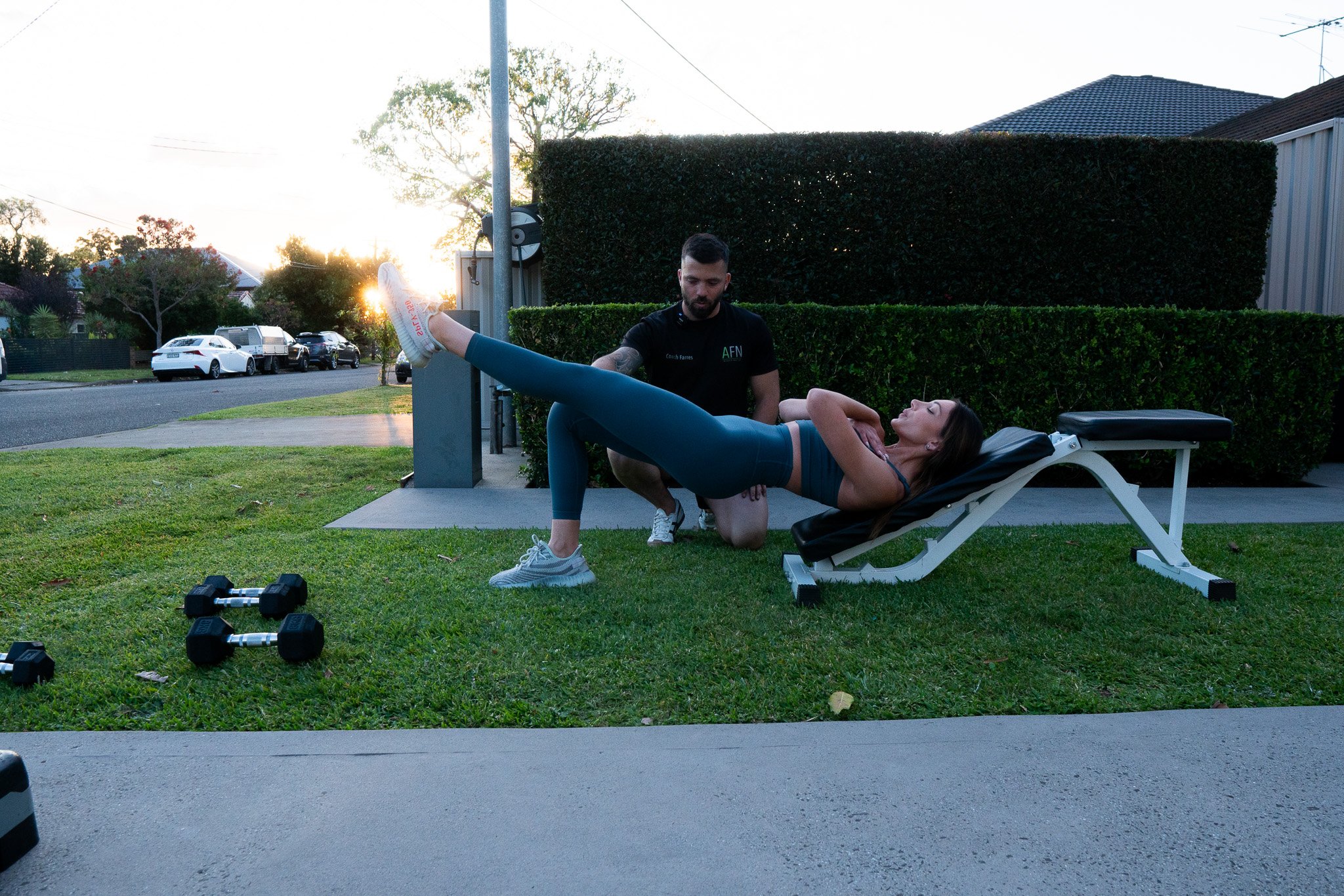 A woman lying on an outdoor massage or therapy table receiving a physical therapy treatment from a man in a black shirt, with some dumbbells on the grass nearby, during sunset in a residential area.