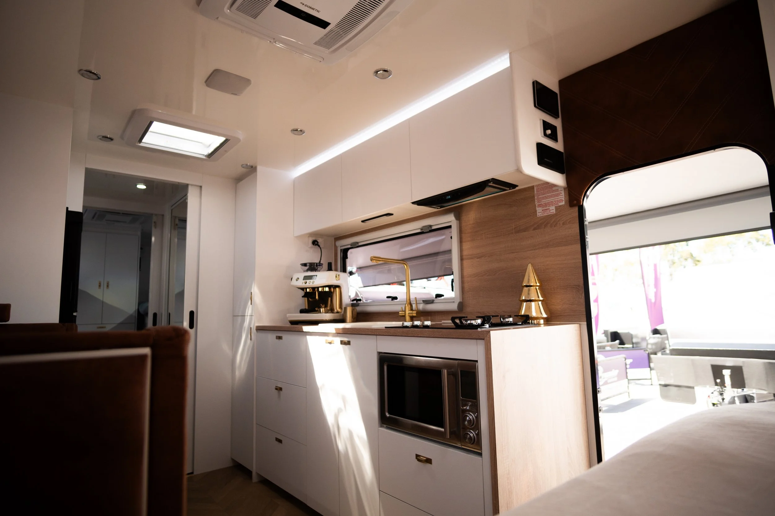Interior of a small, modern kitchen with white cabinetry, a gold faucet, a coffee machine, a microwave, and decorative gold cone-shaped sculptures, with bright natural light coming through small windows.