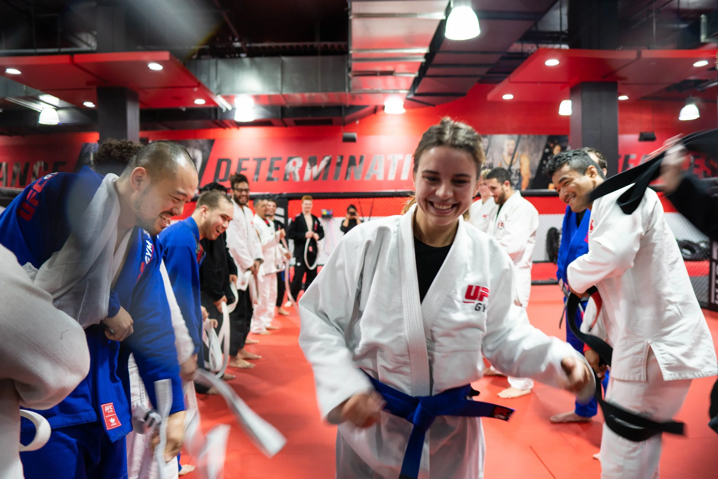 Group of people in martial arts training at UFC gym, smiling and bowing with their belts in a fighting gym with red and black walls, and 'DETERMINATION' written on the wall.